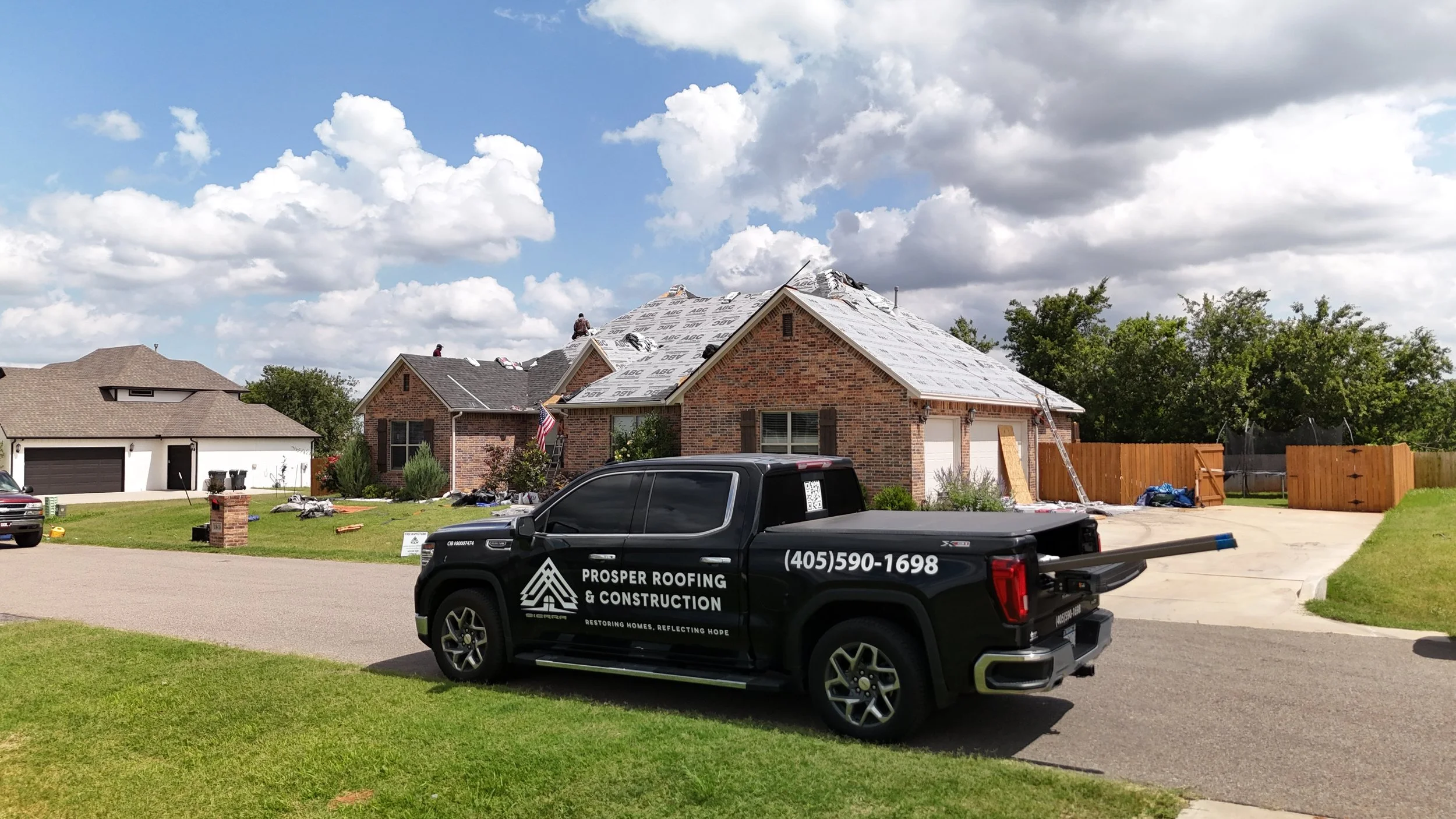 A house under construction with workers on the roof, a black pickup truck in the foreground labeled Prosper Roofing & Construction, and a blue sky with white clouds.