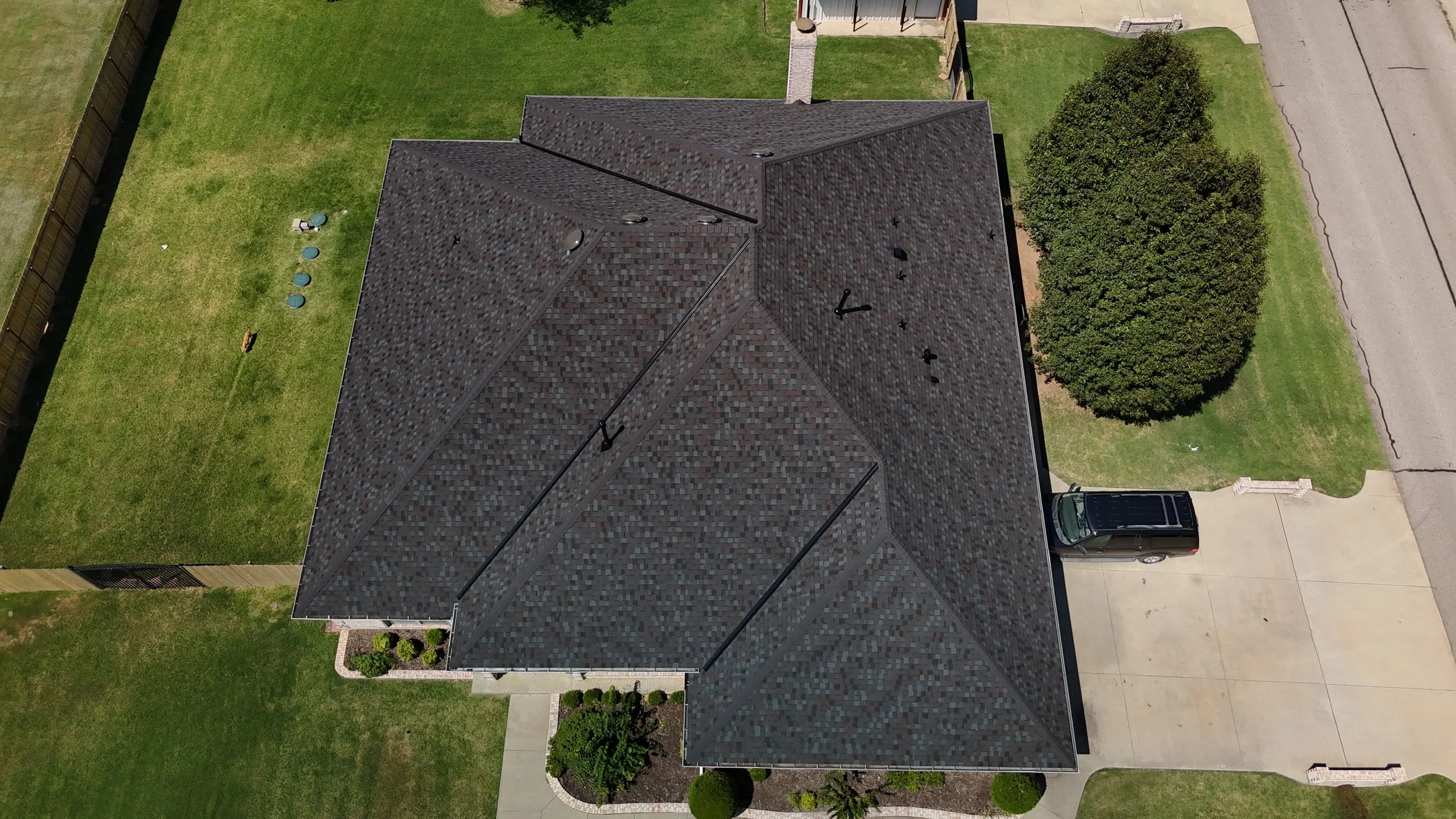 Aerial view of a house with a dark shingle roof, surrounding green yard, large tree, driveway with a vehicle, and small landscaped area at the front.