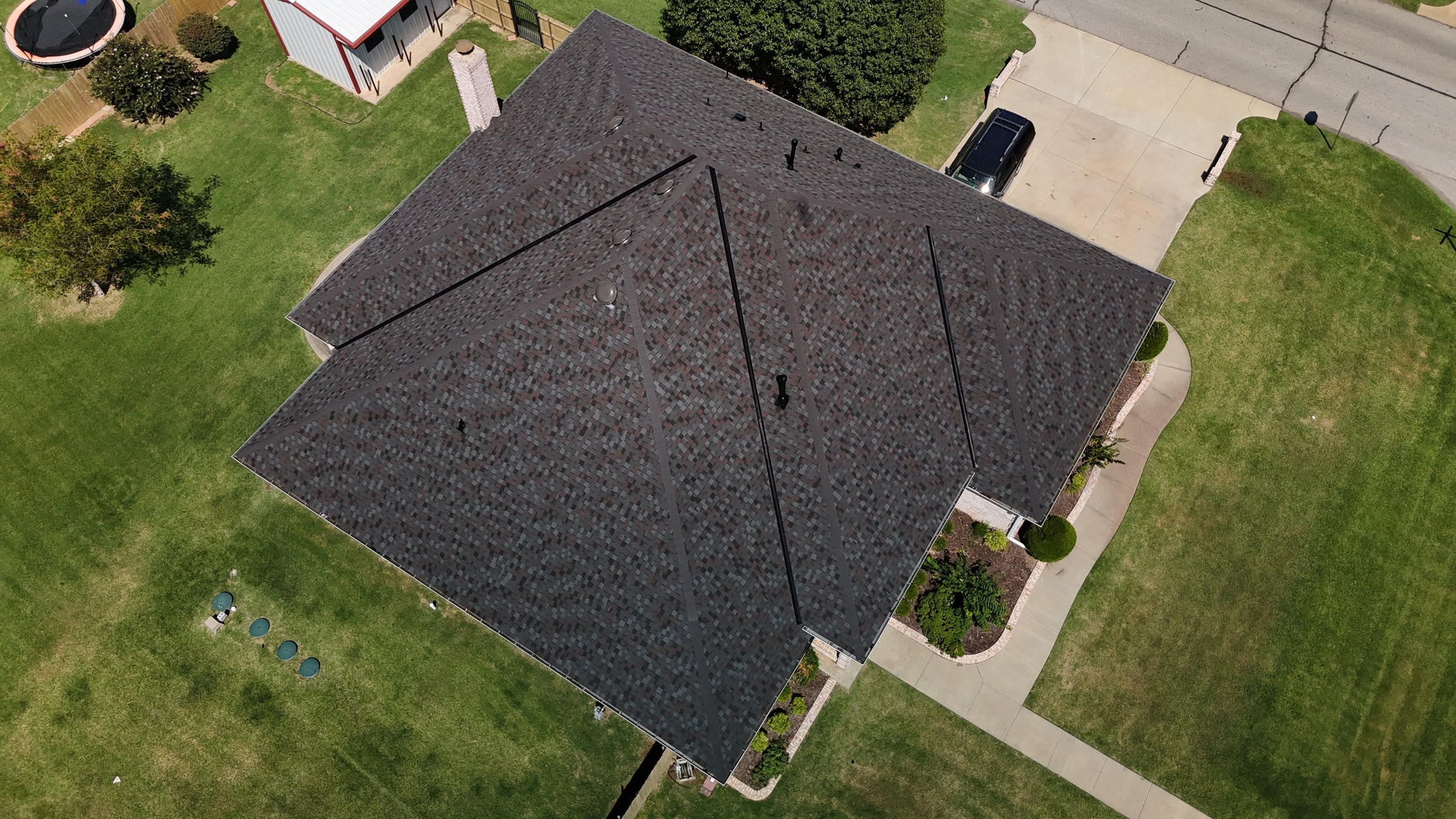 Aerial view of a house with a dark shingled roof, surrounded by a green lawn, trees, and a concrete driveway.