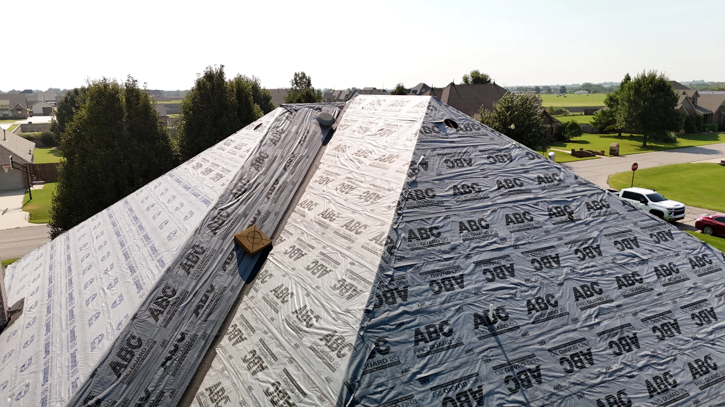 Roof under construction with insulation underlayment, surrounded by residential houses and greenery.