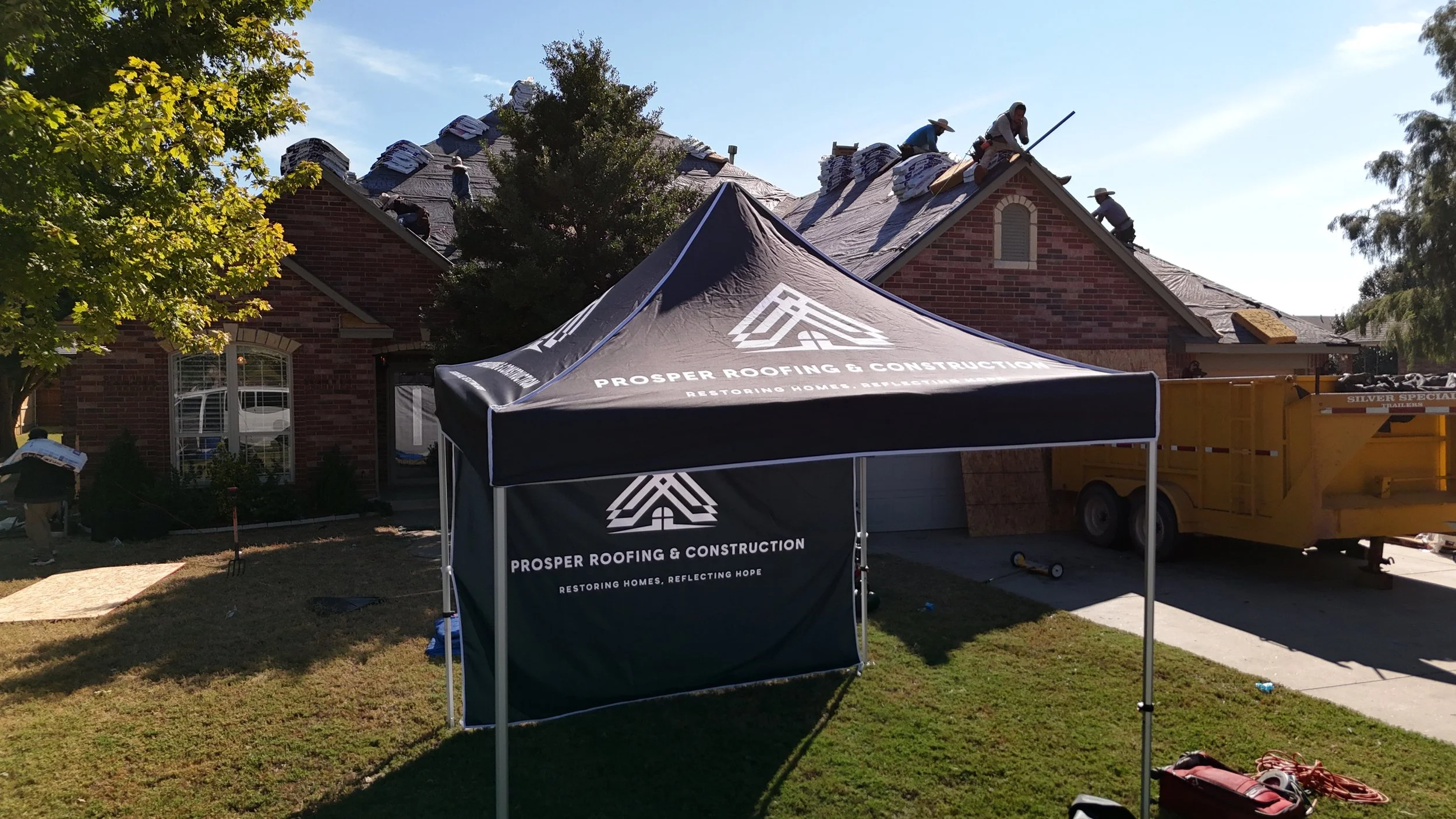 A house under construction with workers on the roof, a black canopy tent with Prosper Roofing & Construction branding in the front yard, and a yellow trailer beside the driveway.