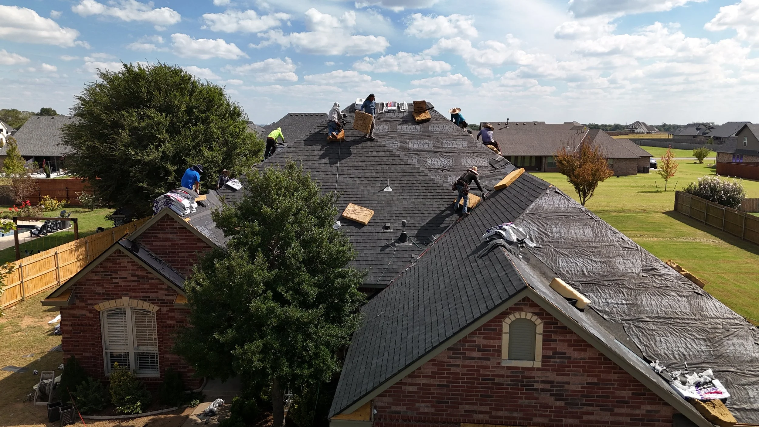 A large group of workers on the roof of a house installing or repairing roofing shingles, with the house surrounded by a wooden fence and trees, under a partly cloudy sky.
