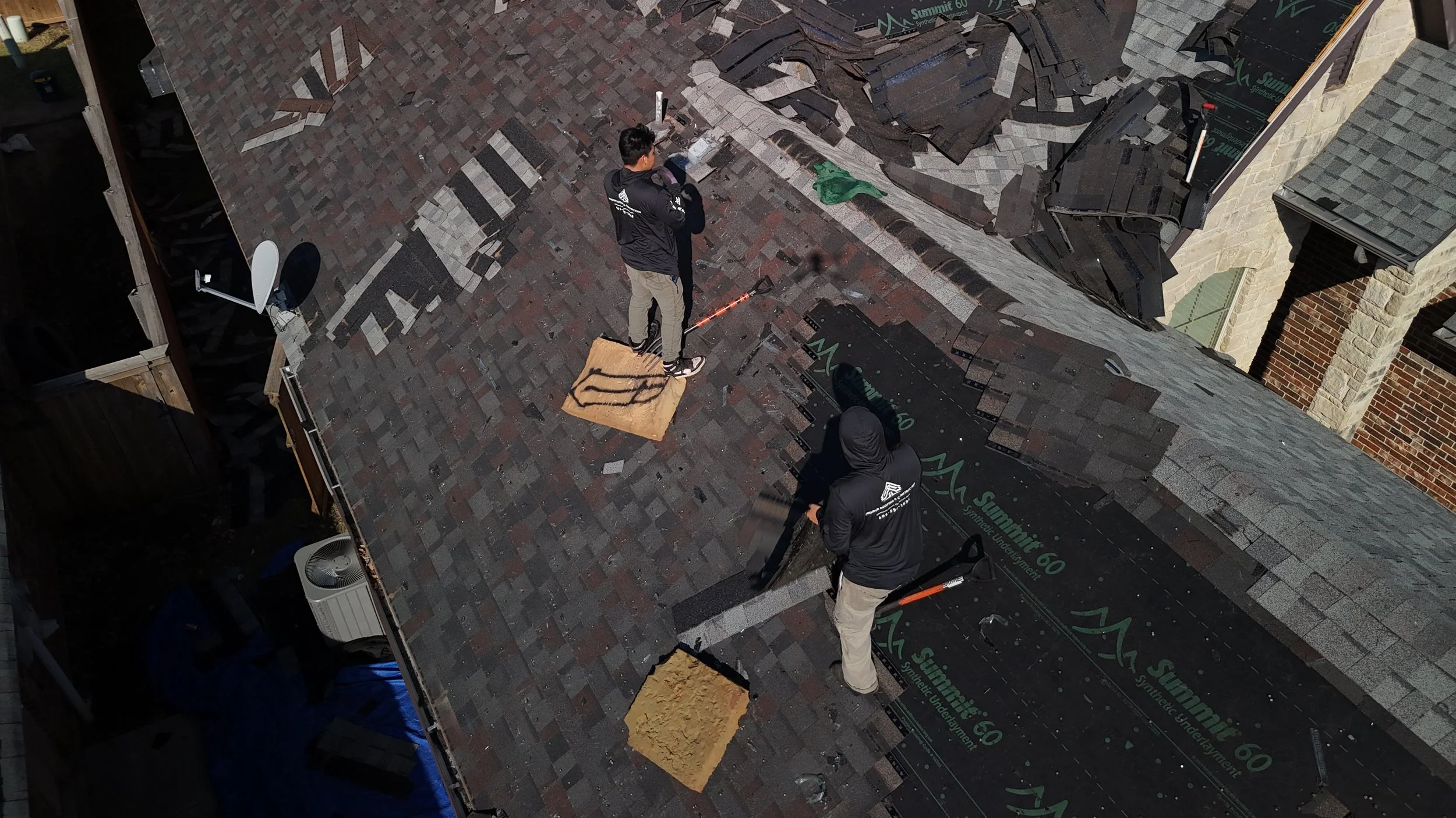 Two workers on a roof installing shingles, with tools and materials around them, some shingles being replaced or repaired.