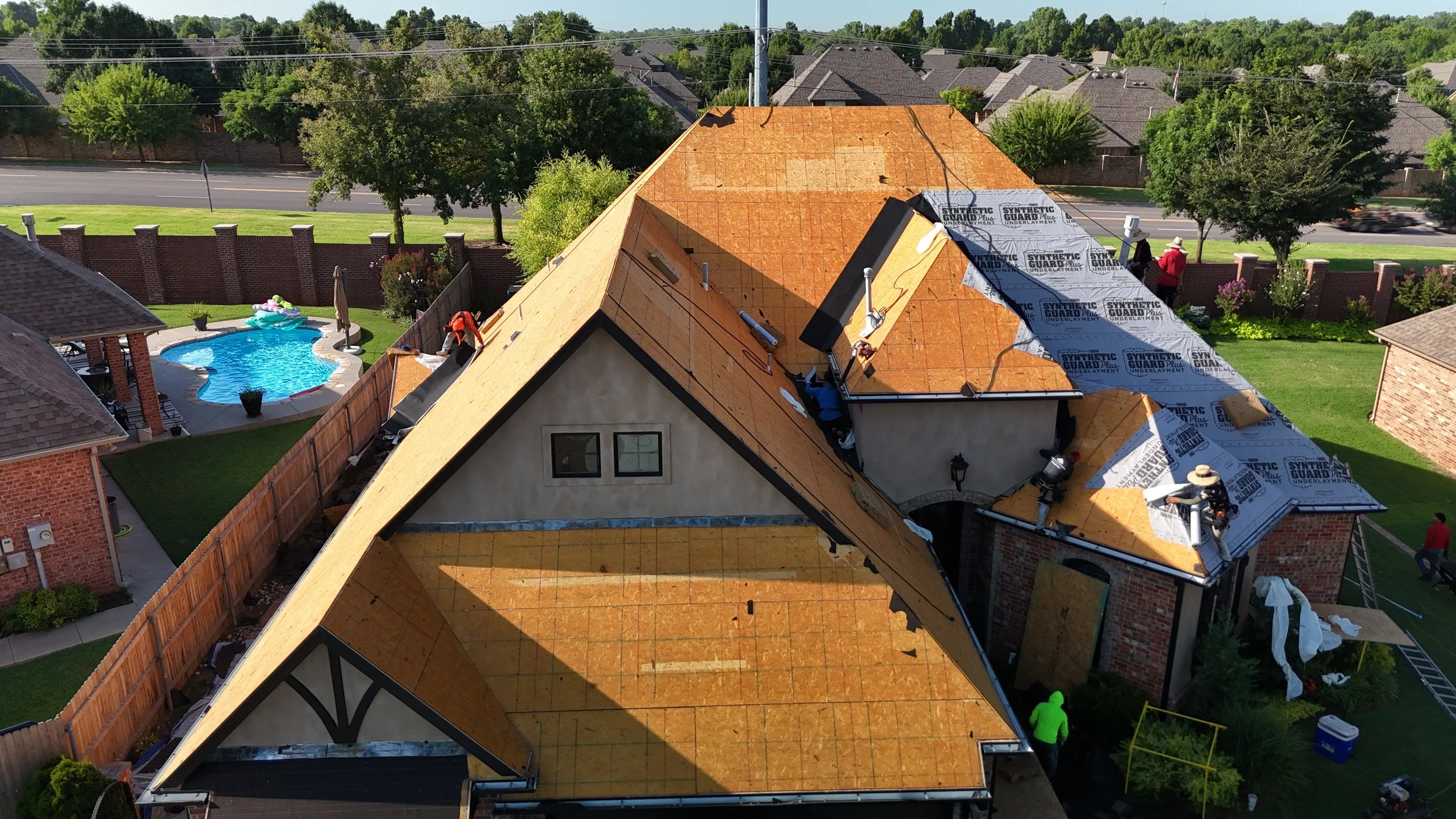 A house under construction with workers installing roofing shingles. The roof is partially covered with protective underlayment. There is a backyard with a pool and a fenced yard, and neighboring houses and a street are visible in the background.