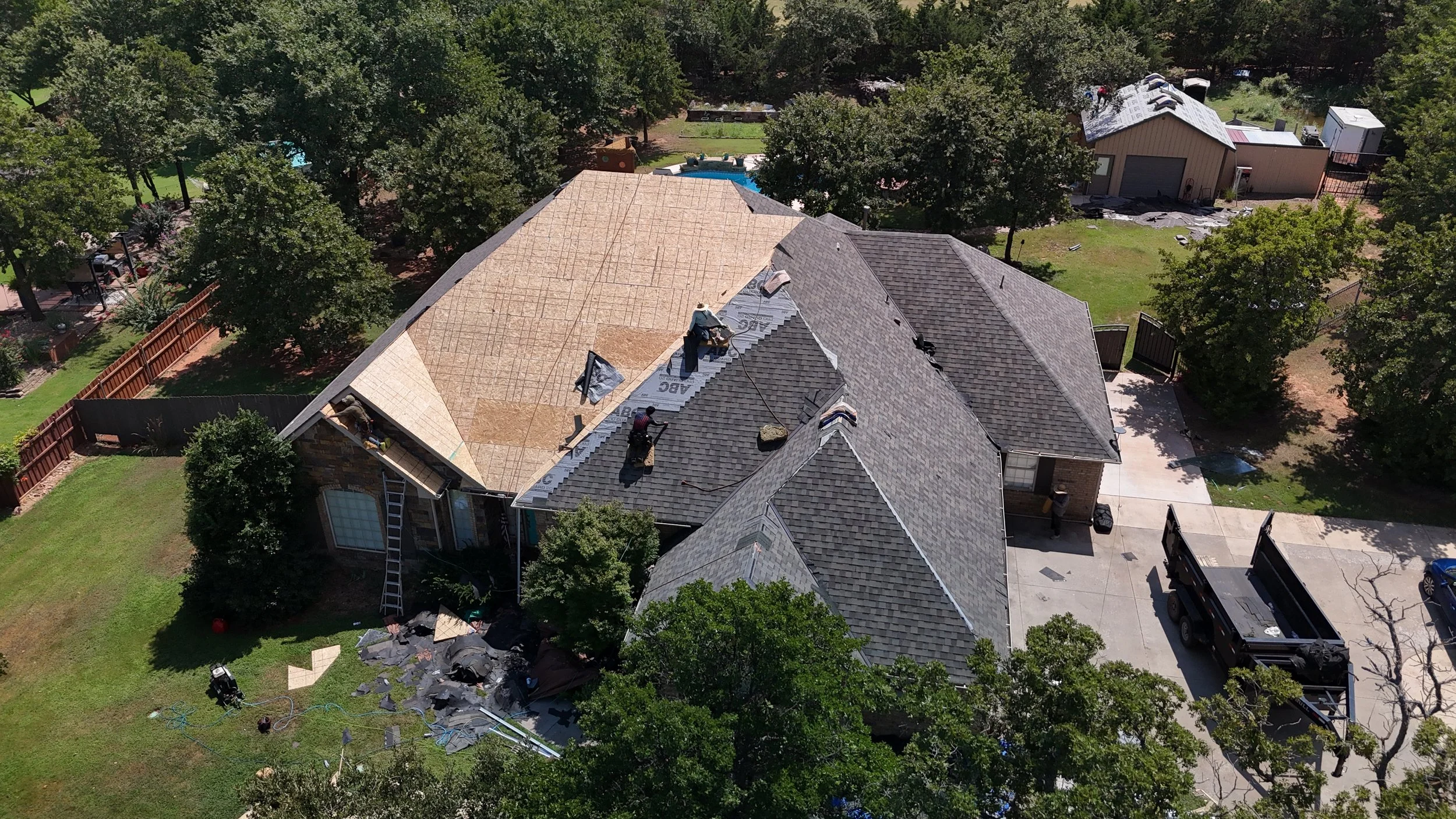 Aerial view of a house under construction with workers installing roofing, construction materials scattered on the lawn, and trees surrounding the property.