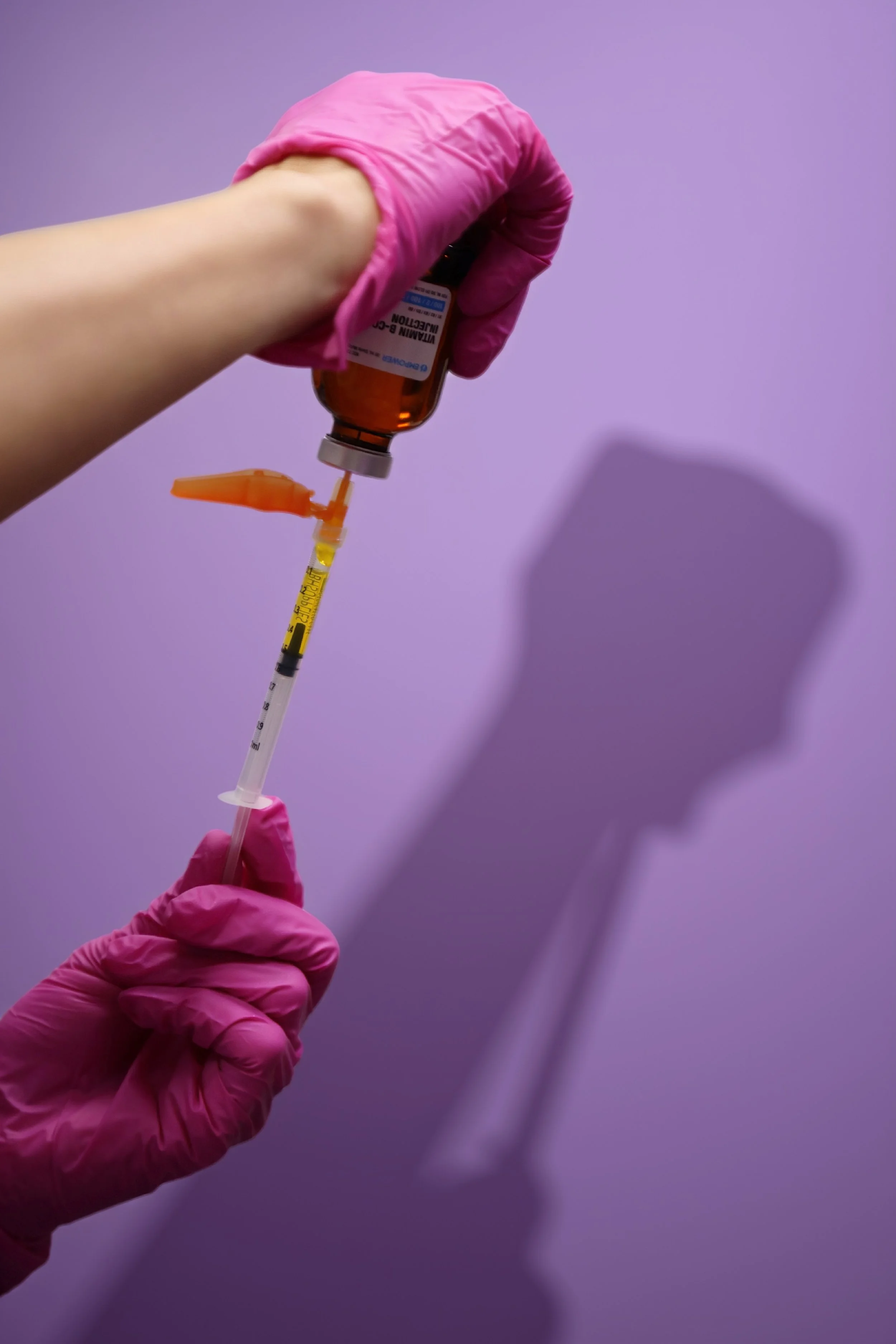 Karie Wilson, RN, BSN, Injector and Owner wearing pink medical gloves; is filling a syringe with 'VITAMIN B-COMPLEX INJECTION'