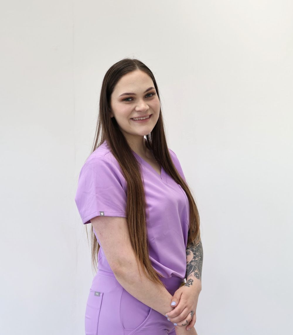 A smiling woman with long, wavy brown hair wearing a black medical uniform, large white feather earrings, and tattoos on her left arm, standing in front of a floral mural background.