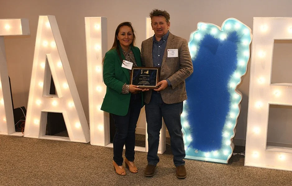 Two people holding an award plaque in front of lighted letters and a decorative backdrop.