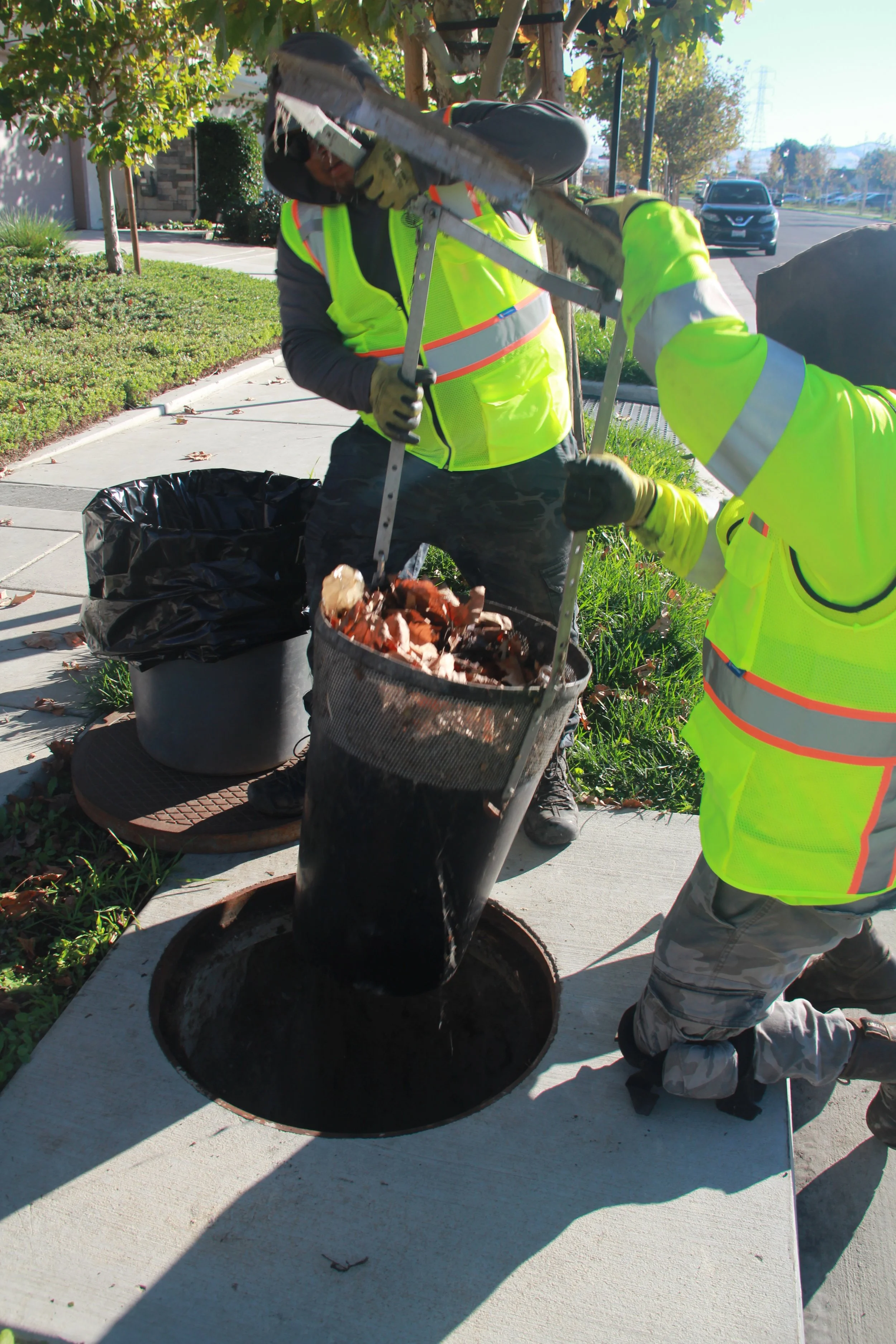 Workers in yellow safety vests and gloves using a tool to remove a manhole cover on a sidewalk.