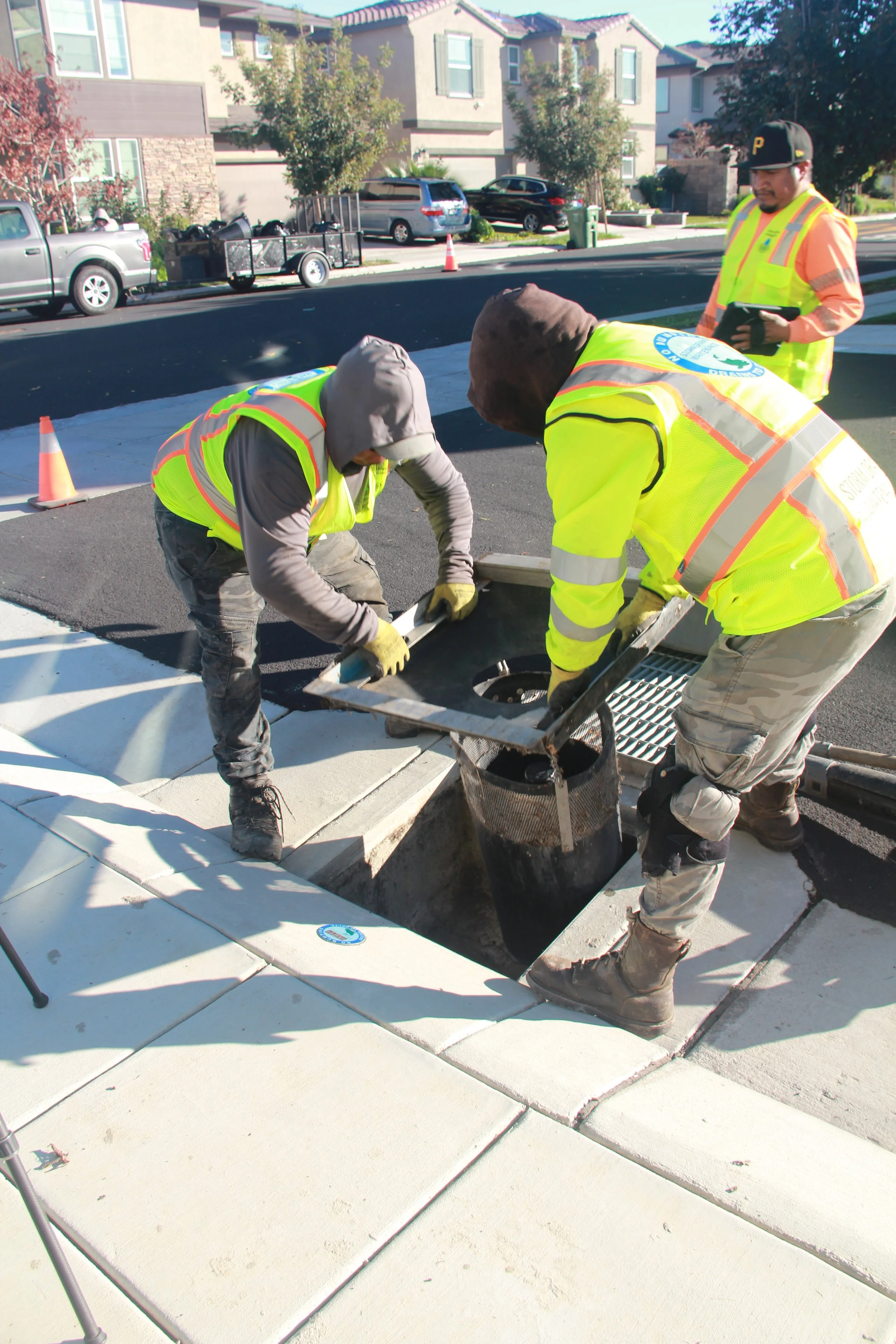 Water Decontaminator installation team workers installing a storm drain catch basin filter on a city sidewalk, with orange cones and parked cars in the background.