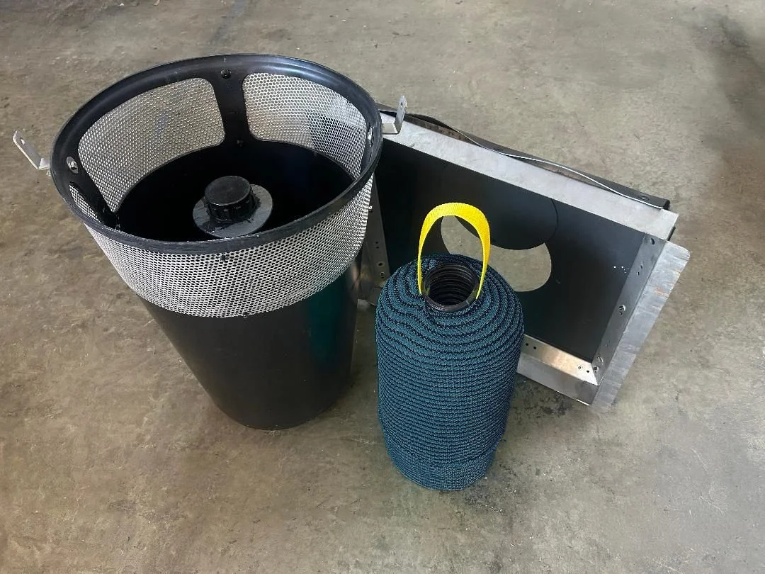 A full trash capture device with a mesh outer shell, a black cylindrical bucket, and a separate filter with a yellow handle, arranged on a concrete floor.