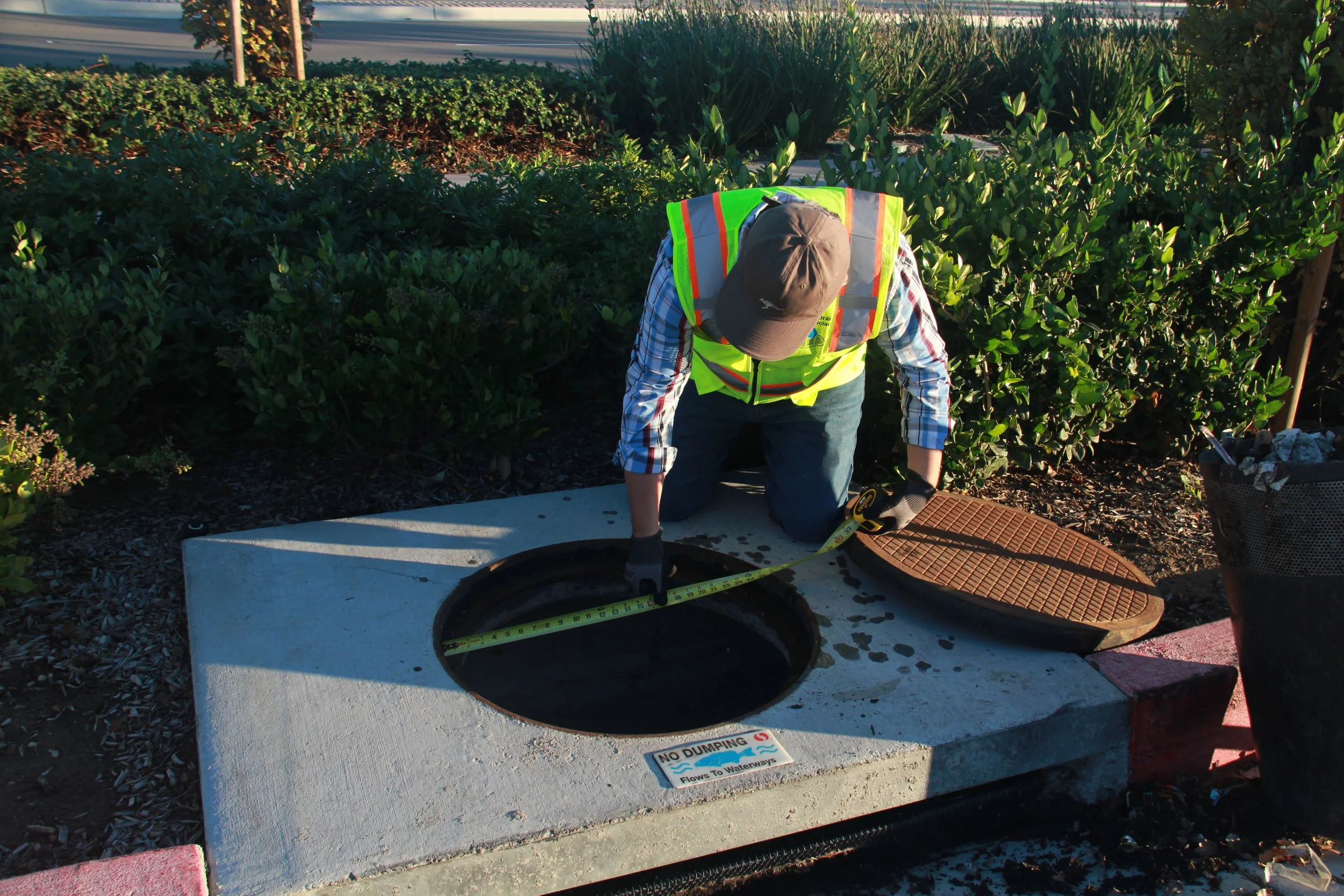 A worker in a safety vest and gloves is checking the diameter of a storm drain with a measuring tape in an outdoor setting with bushes and a sidewalk.