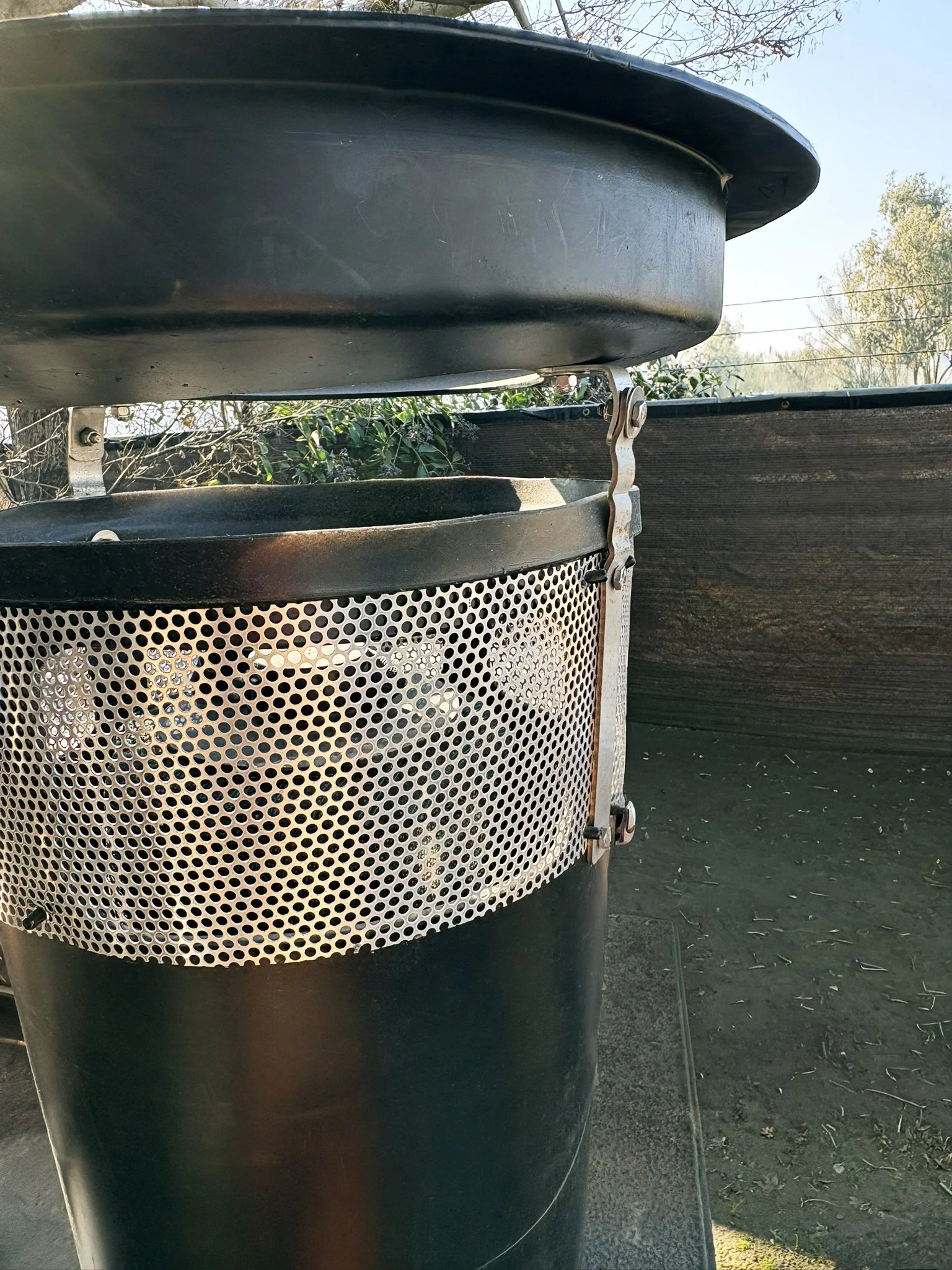 Close-up of aa Water Decontaminator Full Trash Capture Catch Basin Insert, including a black cylindrical catch bucket with a metal mesh, outdoors with trees and a blue sky in the background.