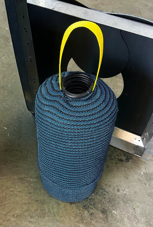 A filter for the Water Decontaminator Trash Full Capture device with a yellow handle, standing on a concrete floor next to a black metal storm drain collar.