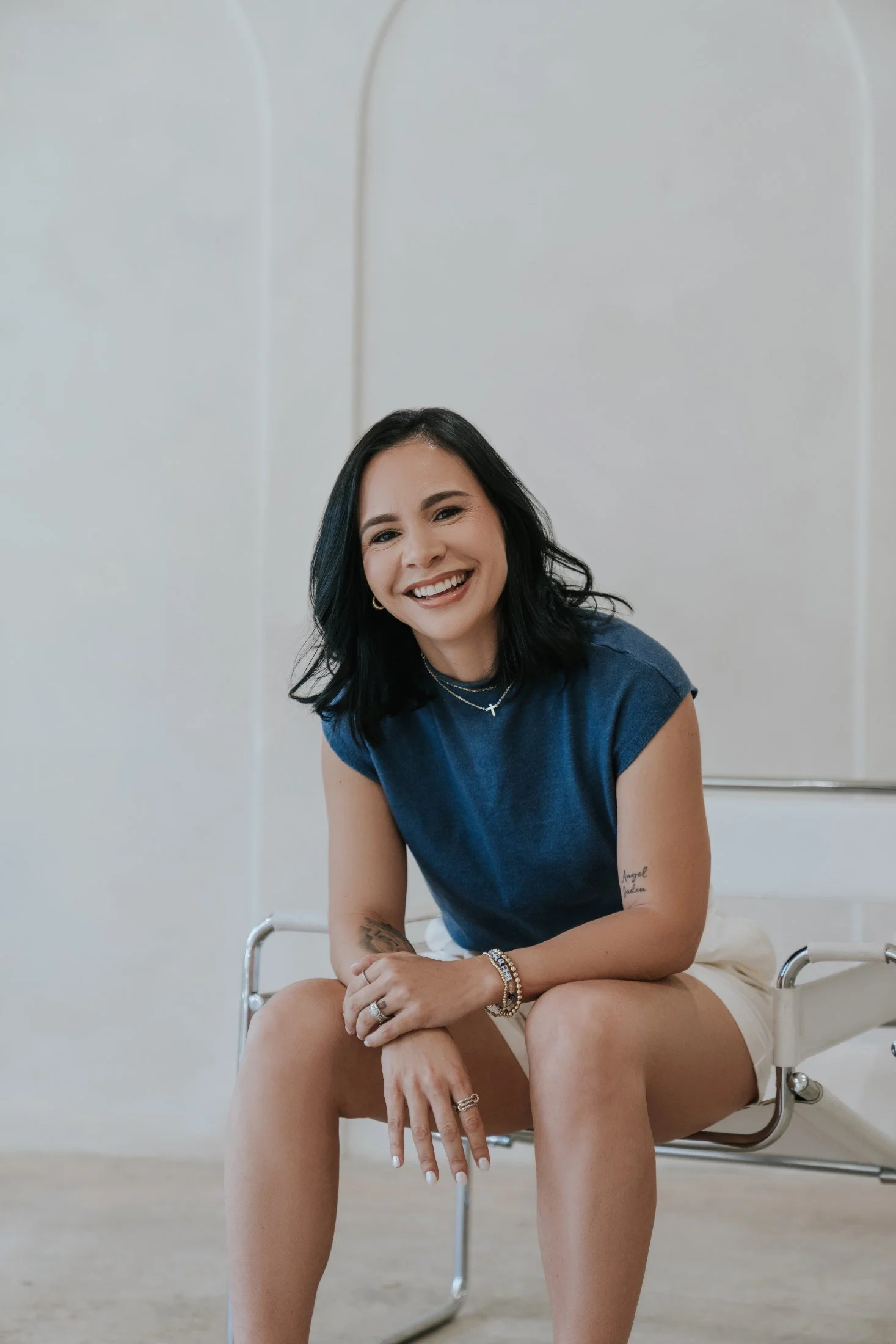 A woman with long dark hair smiling and sitting behind a white cushioned object on a wooden stand against a plain white background.