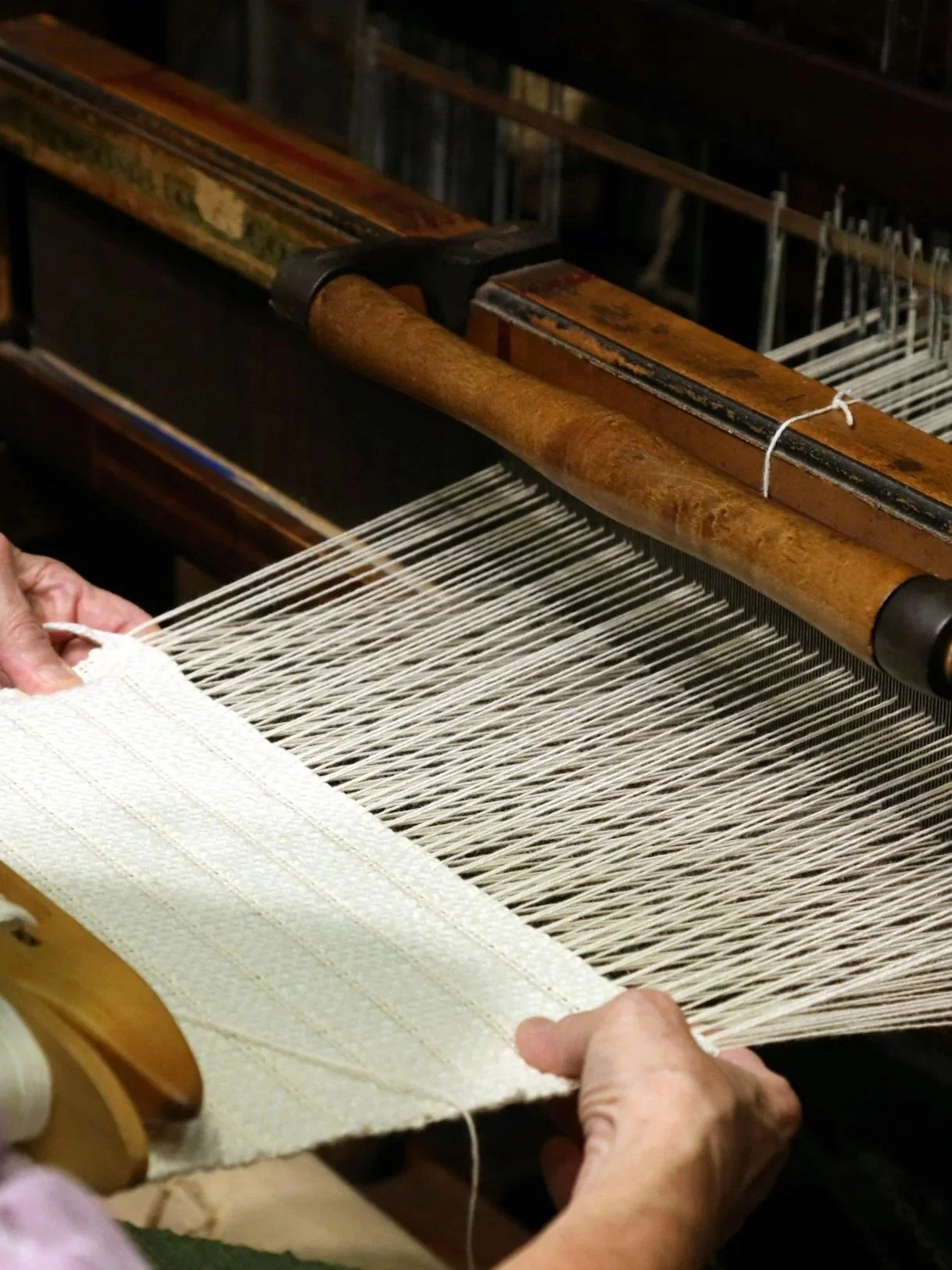 Hands working on a traditional loom, weaving white fabric with multiple threads, in a craft or textile workshop.
