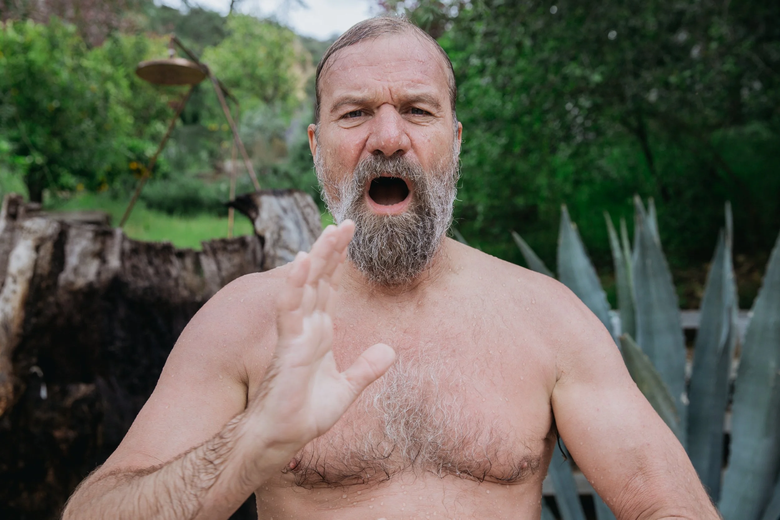 Man with a beard and bare chest outdoors, with mouth open and hand raised, surrounded by greenery.