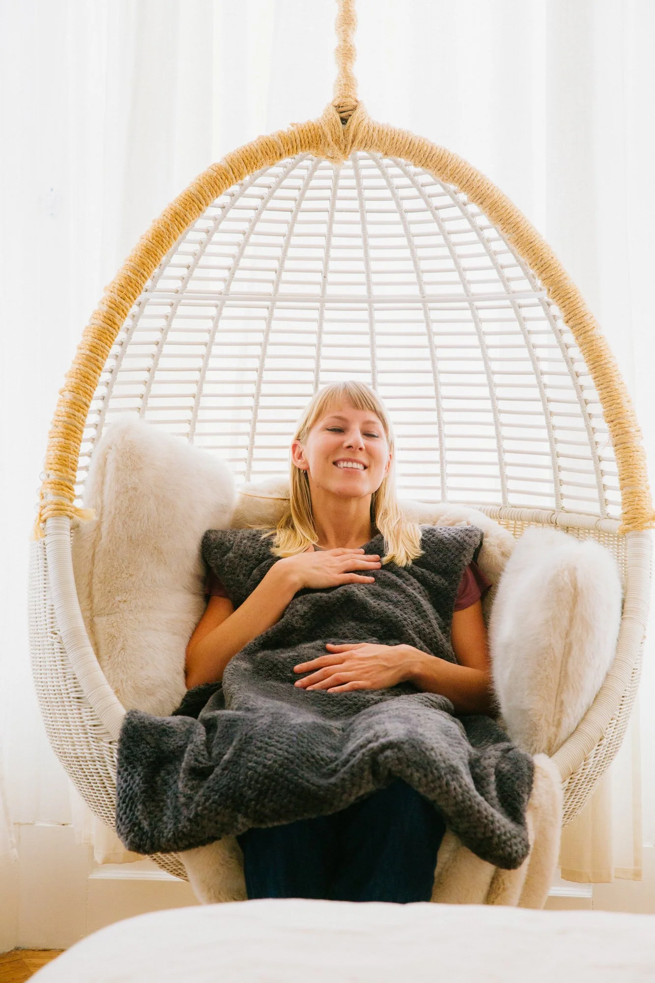 Person relaxing in a hanging wicker chair with a cozy blanket in a bright room.