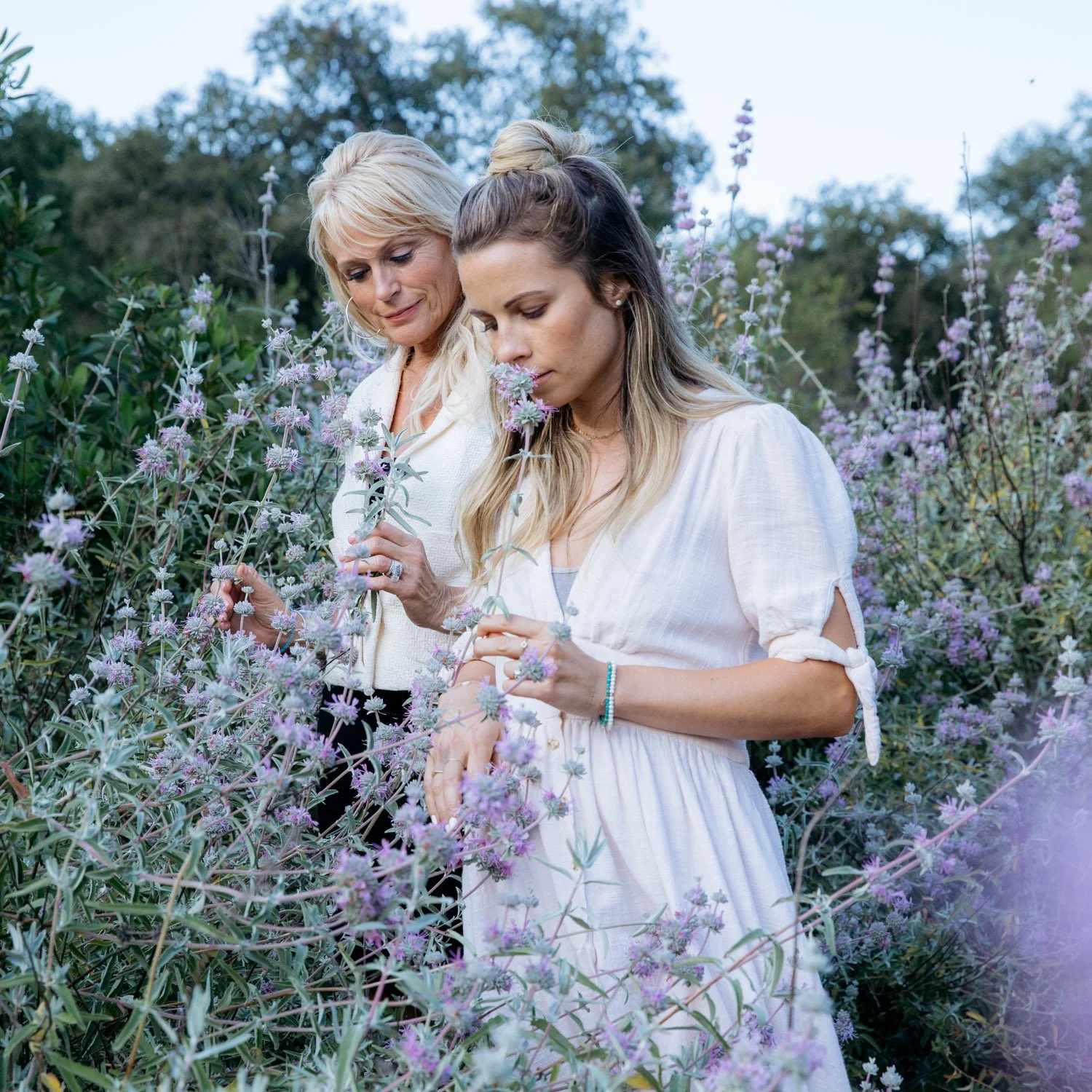 Two women in a field of purple flowers, one smelling a flower, both wearing light-colored dresses.