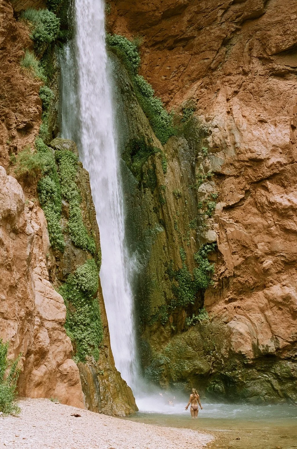 Waterfall cascading down a rocky cliff with greenery, and a person standing in a pool at the base.