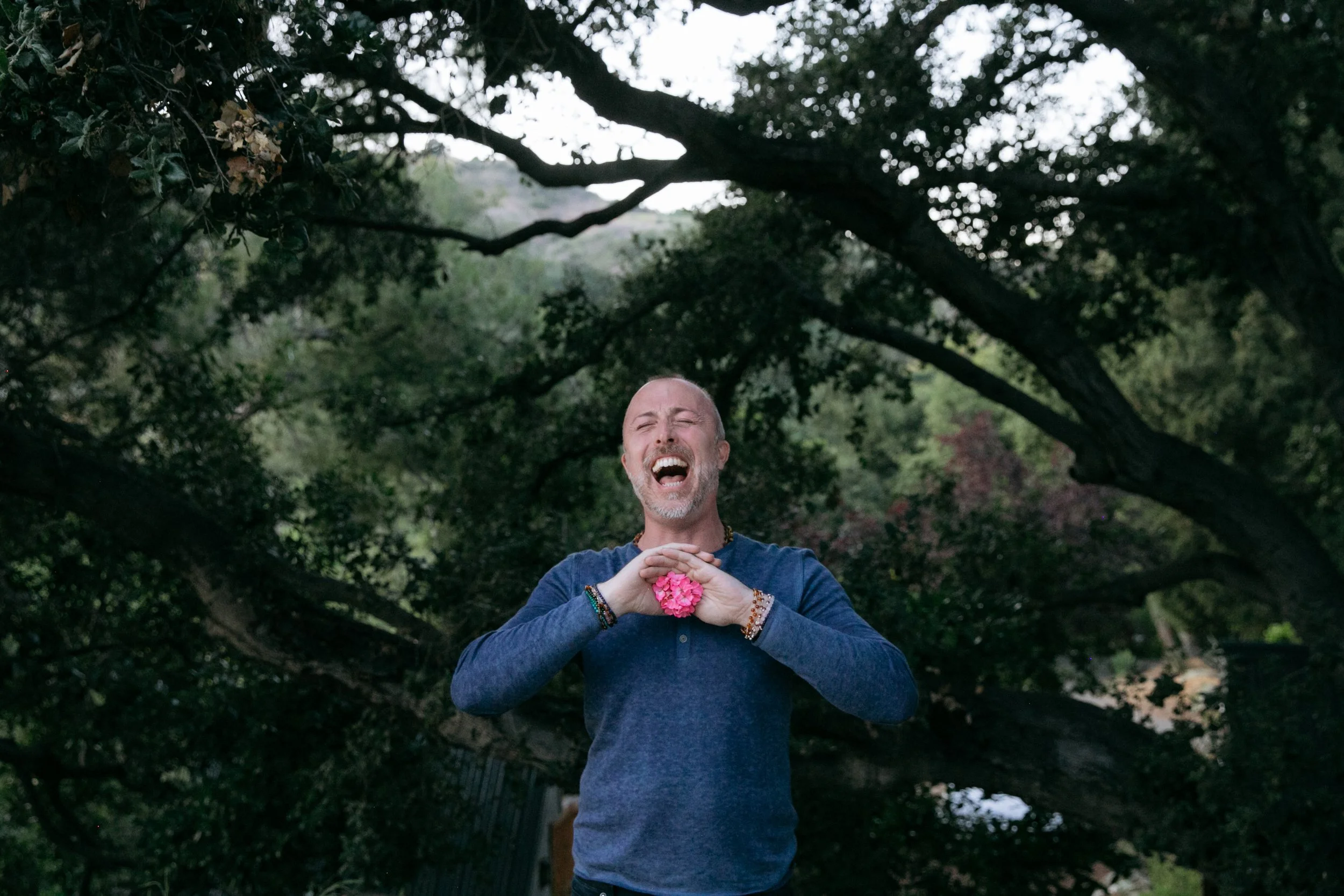 Person outdoors laughing and holding a pink flower under trees.