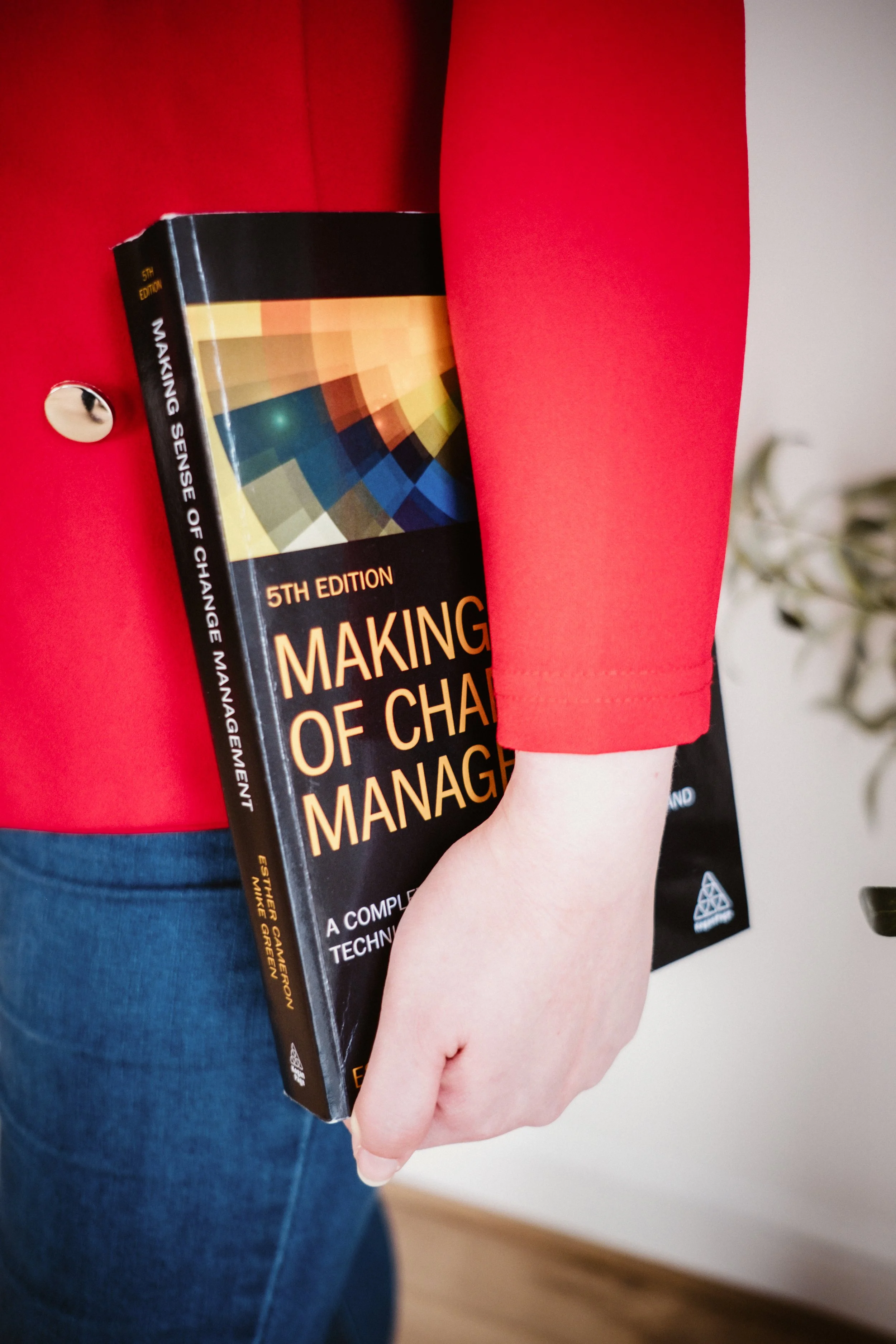 Side profile of a woman's hip. She's wearing blue jeans and a red blazer. She holds a book on change management against her hip