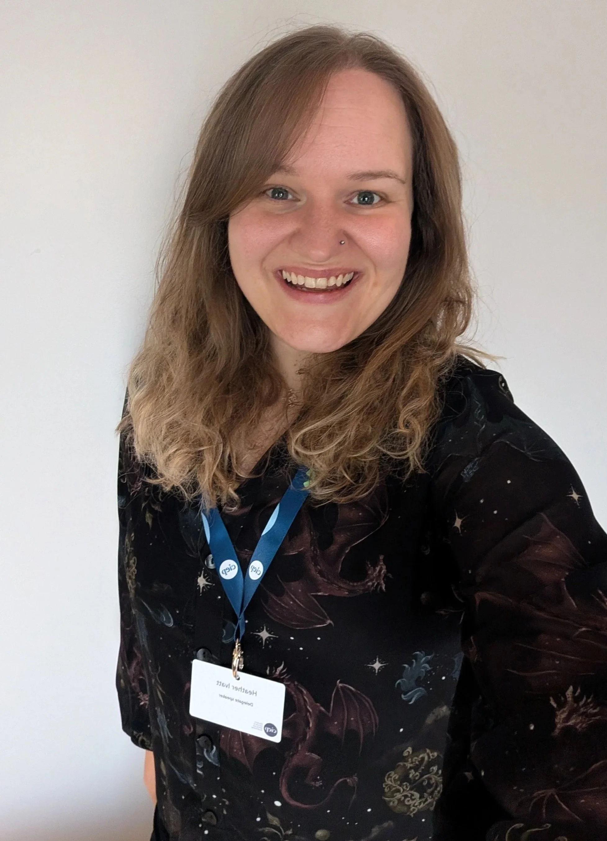 A woman with blonde hair smiles at the camera as she takes a selfie. She is wearing a fantasy-themed shirt that has images of dragons and stars on it. A CIEP lanyard is around her neck, holding a name badge.