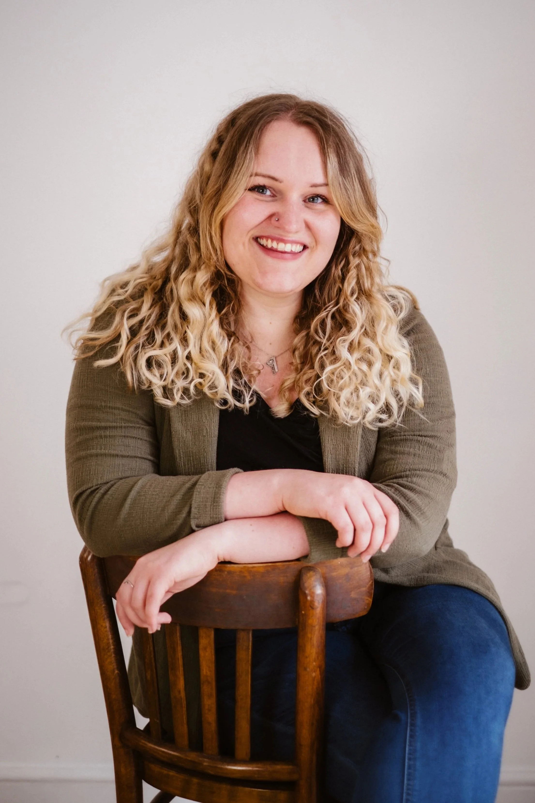 A woman with long curly blonde hair sitting on a wooden chair, smiling, wearing a green blazer and black shirt, against a plain white wall.