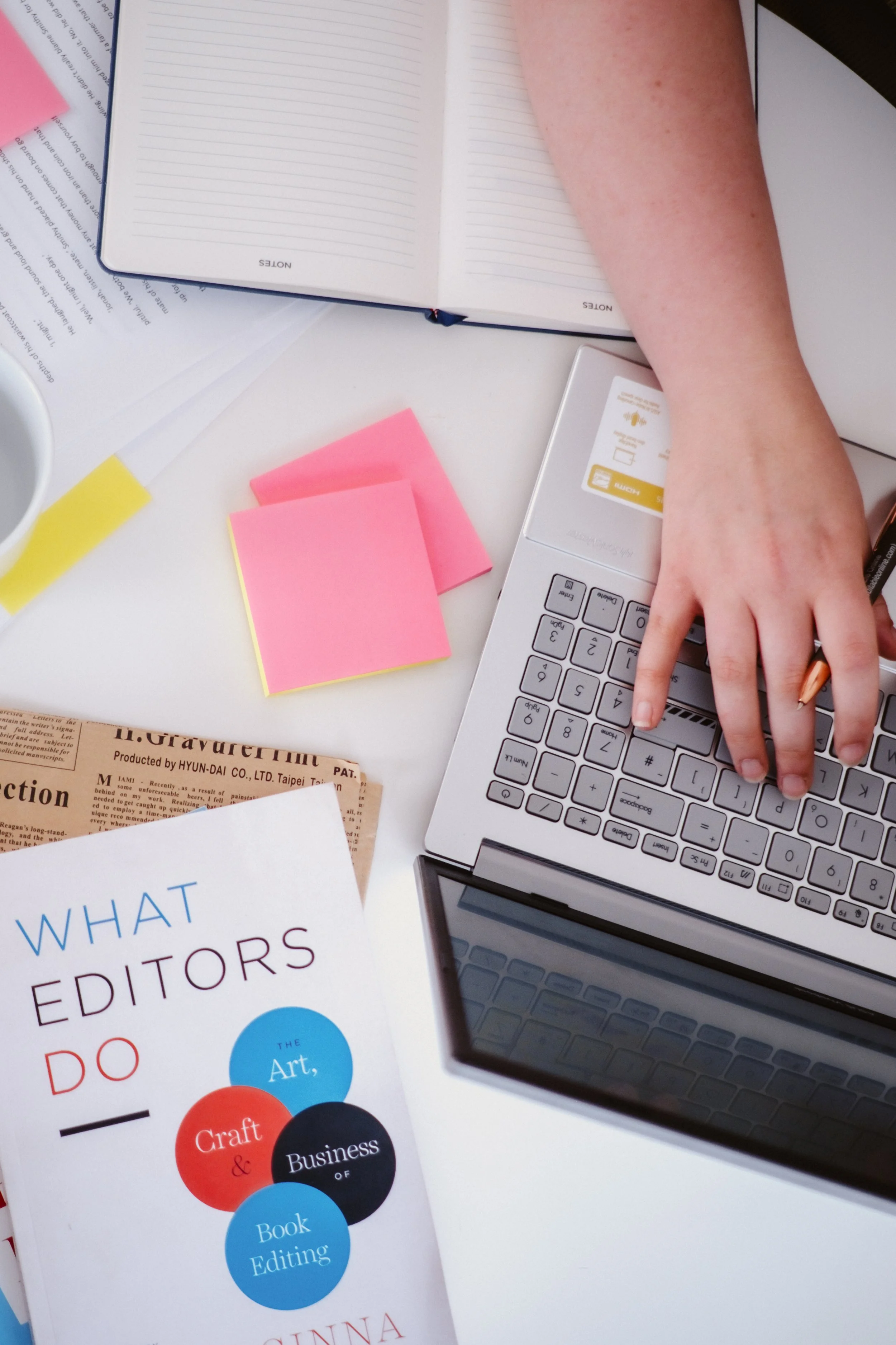 Image of a desk with sticky notes, newspaper, and a laptop keyboard. A woman's hand is visible holding a pen as she types on the keyboard.
