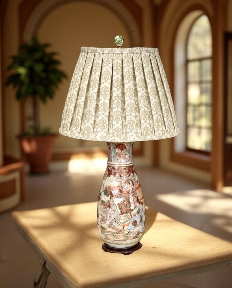 Table lamp with a patterned ceramic base and a pleated fabric lampshade, on a wooden table inside a room with large arched windows.