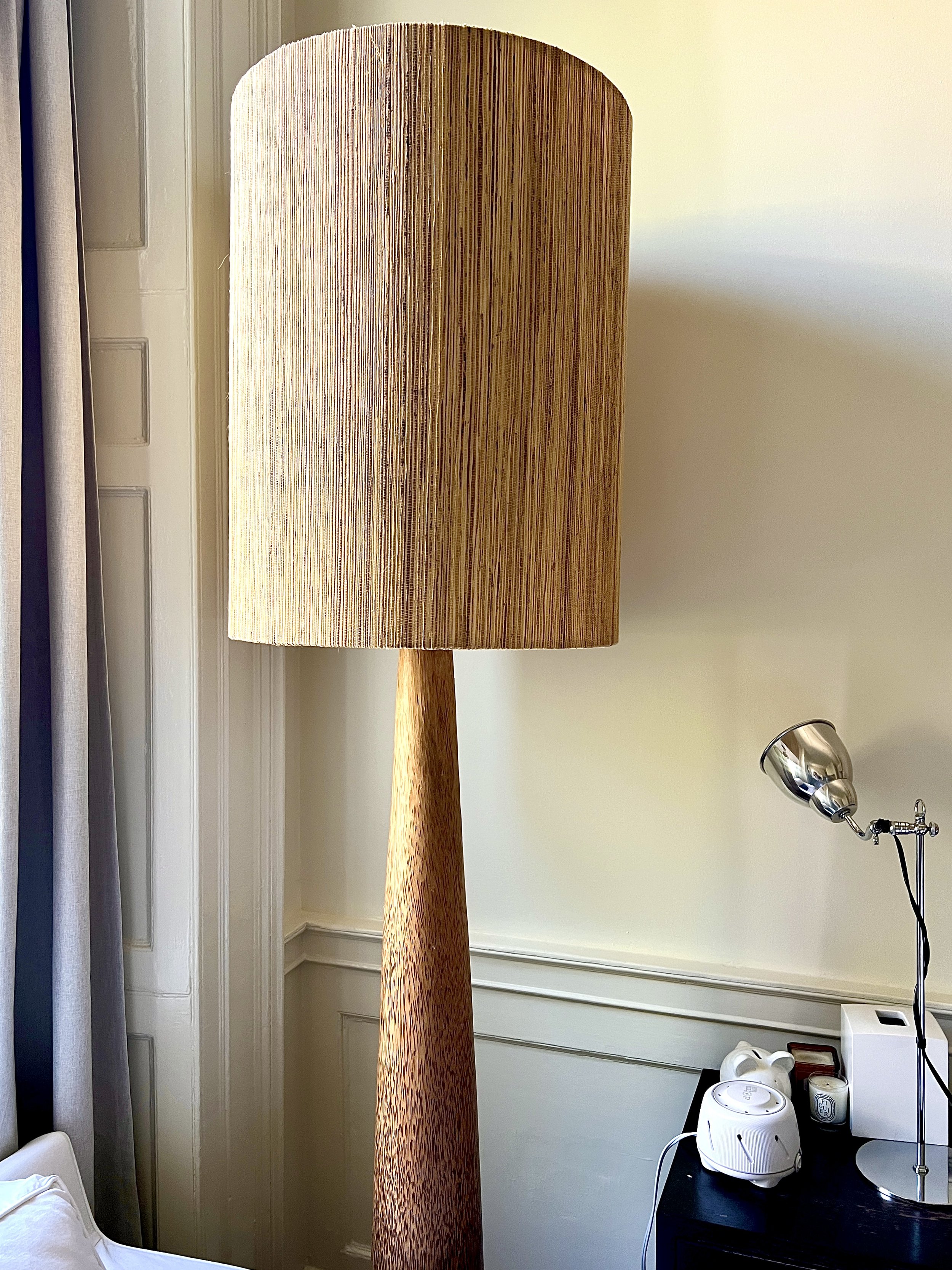 A tall wooden floor lamp with a vertical striped fabric shade standing next to a wall in a room with a side table that holds a white humidifier, a small clock, and a metallic desk lamp.