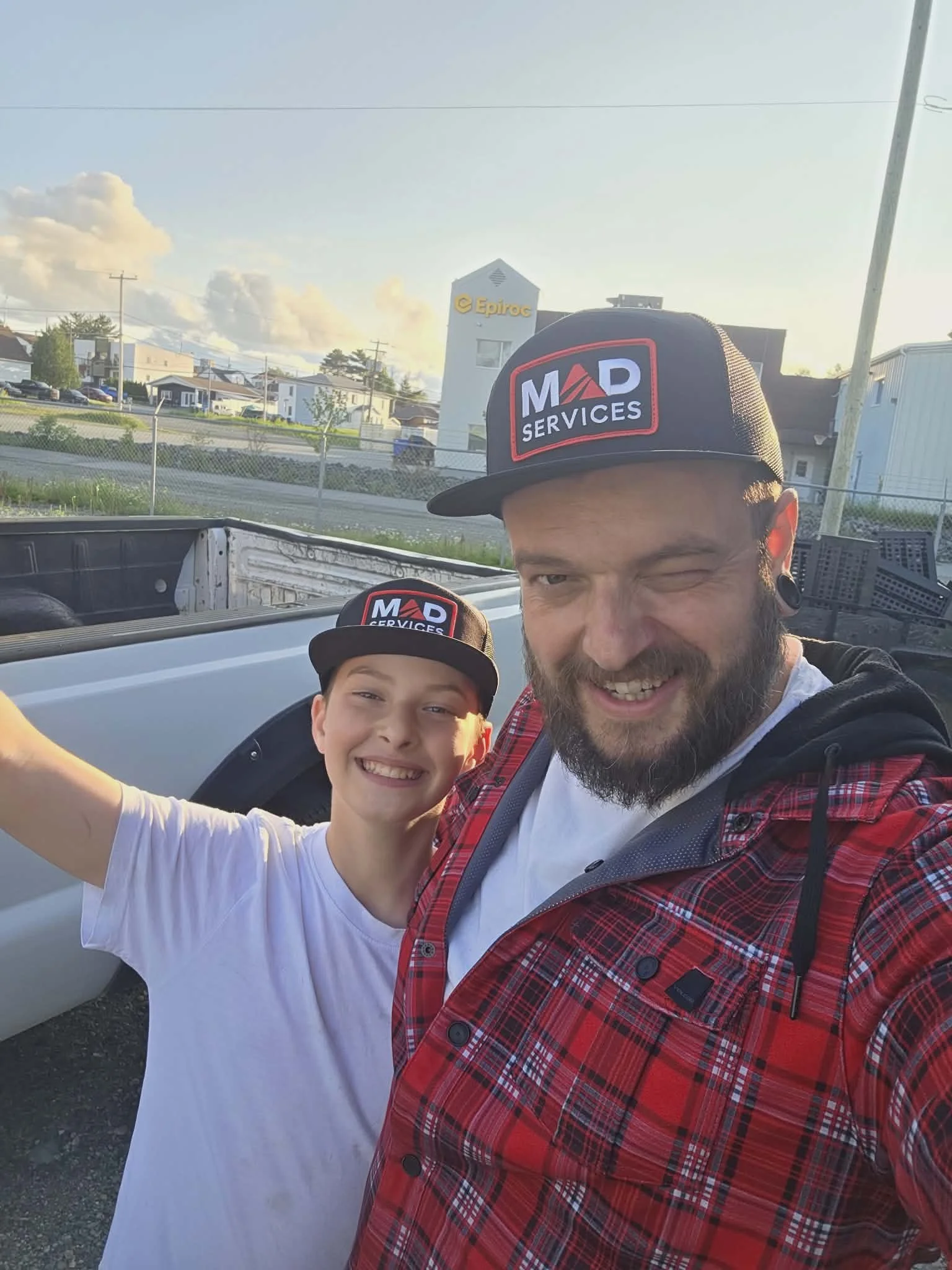 Deux personnes souriantes posant pour une photo à l'extérieur, portant des casquettes avec le logo 'MAD Services'.
