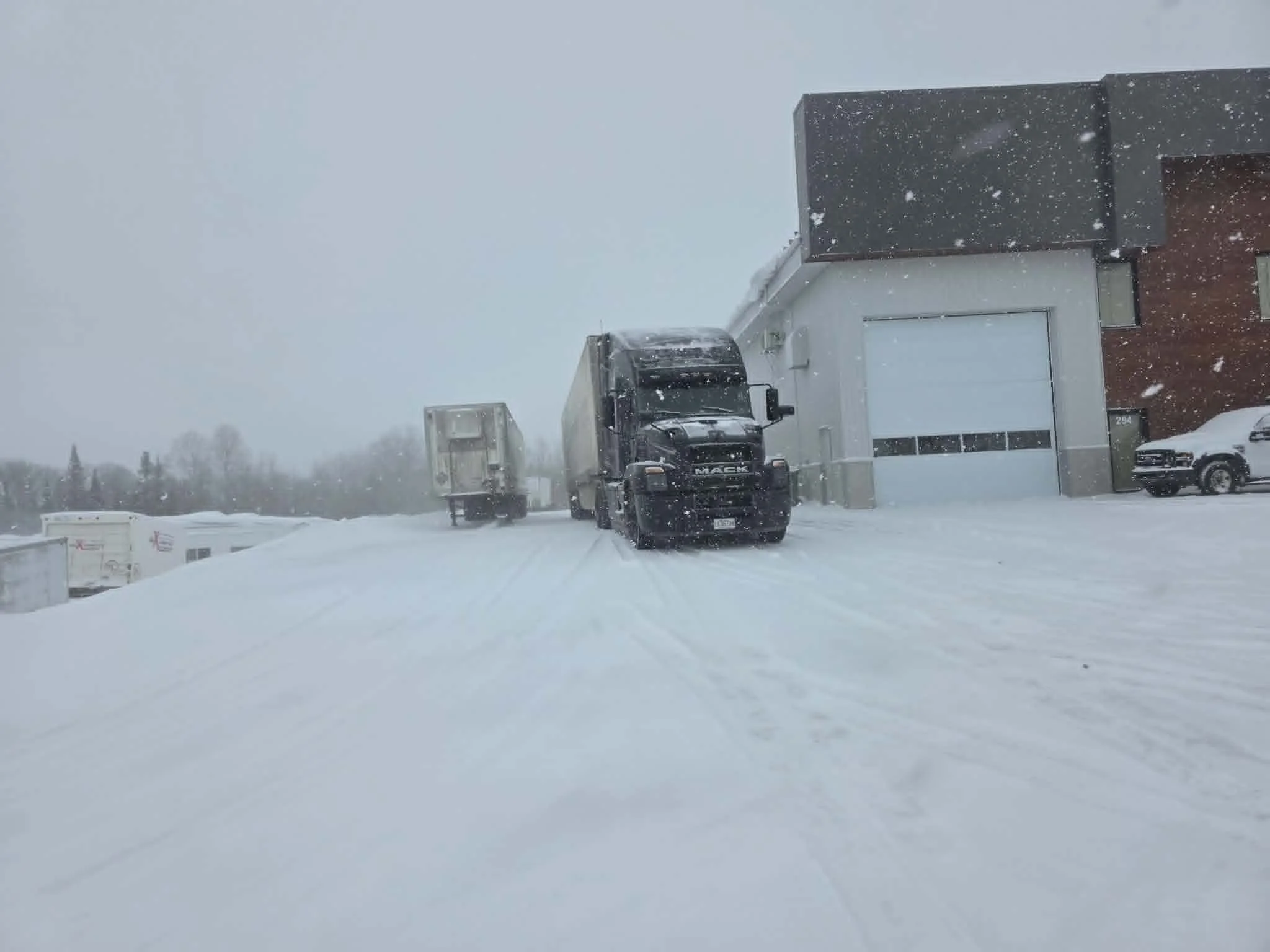 Un bâtiment moderne avec un garage, un camion noir et une voiture blanche garés devant, sous une forte chute de neige.