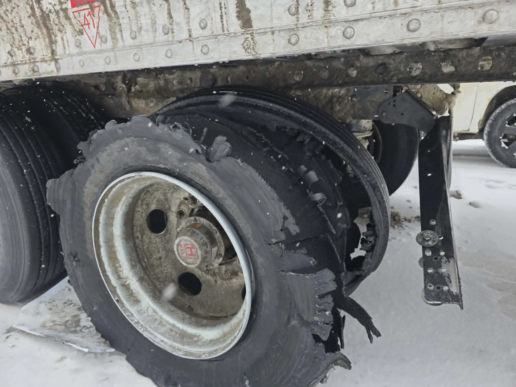 Un camion est endommagé, avec la roue arrière dégonflée ou cassée, sur fond de neige, avec une petite chienne visible à l'arrière du camion.
