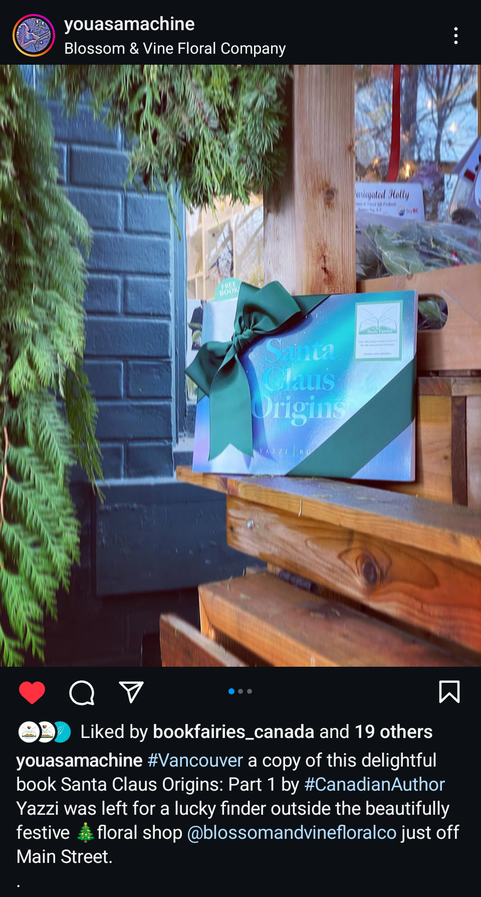 A festive display with a blue sign reading "Santa Claus Origins" decorated with a green ribbon bow, placed on a wooden table outside a floral shop with greenery and a brick wall in the background.