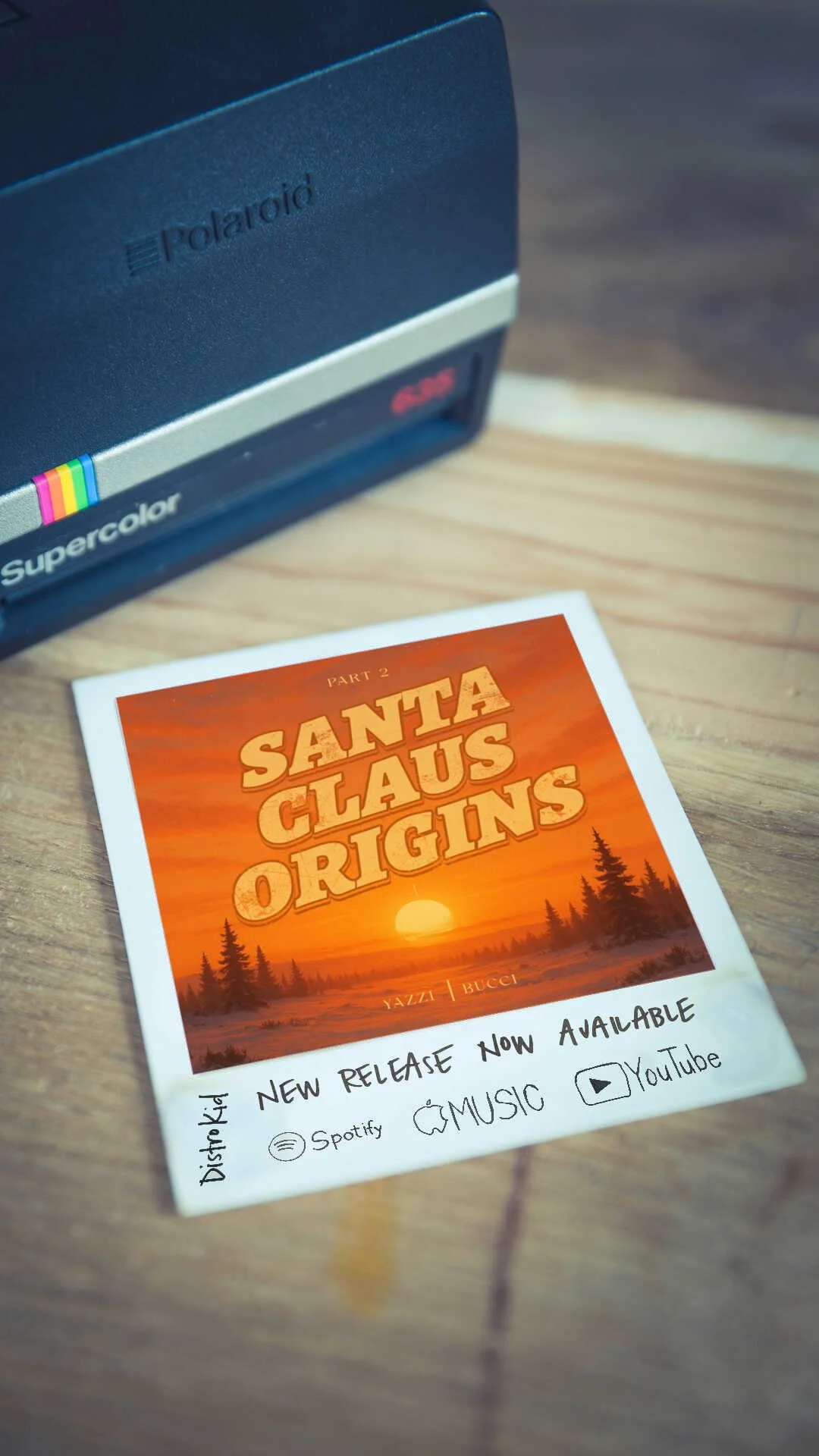 A Polaroid-style photo of a sunset scene with trees and the text "Santa Claus Origins" on top, advertising a new release available on Spotify, Apple Music, and YouTube, placed on a wooden surface near a polaroid camera.