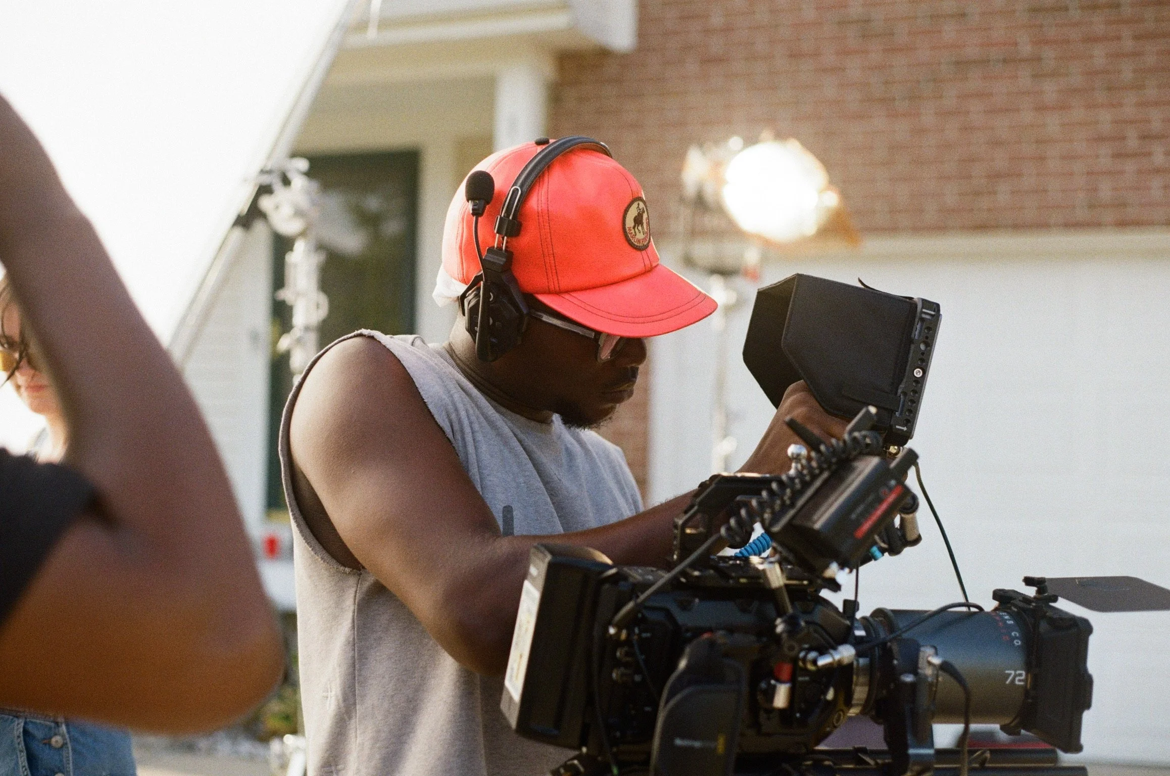 A man wearing a red cap and black headphones operating a professional video camera outdoors, with a woman partially visible in the background.