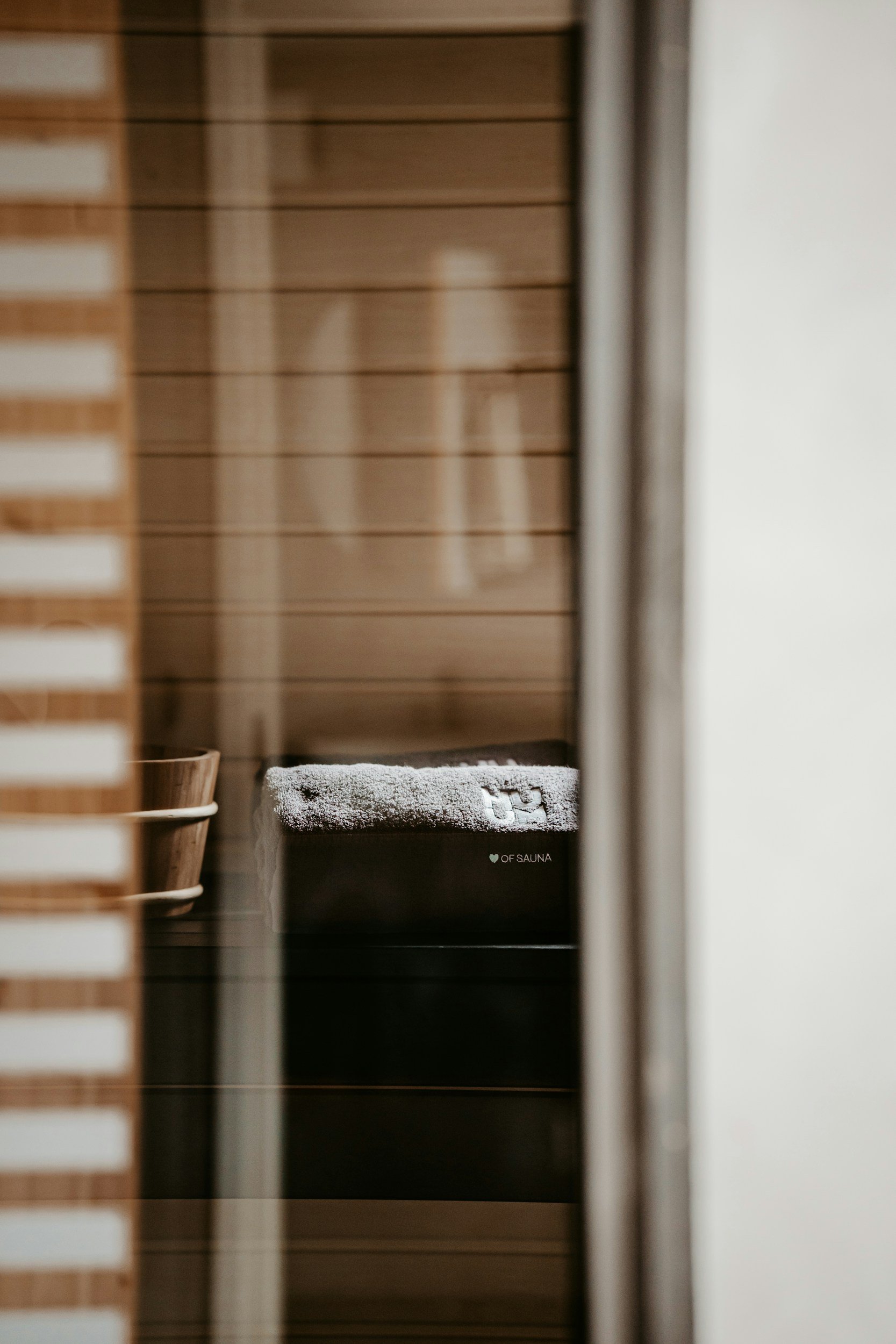 View through a window showing a wooden sauna interior with a towel and wooden bucket inside.