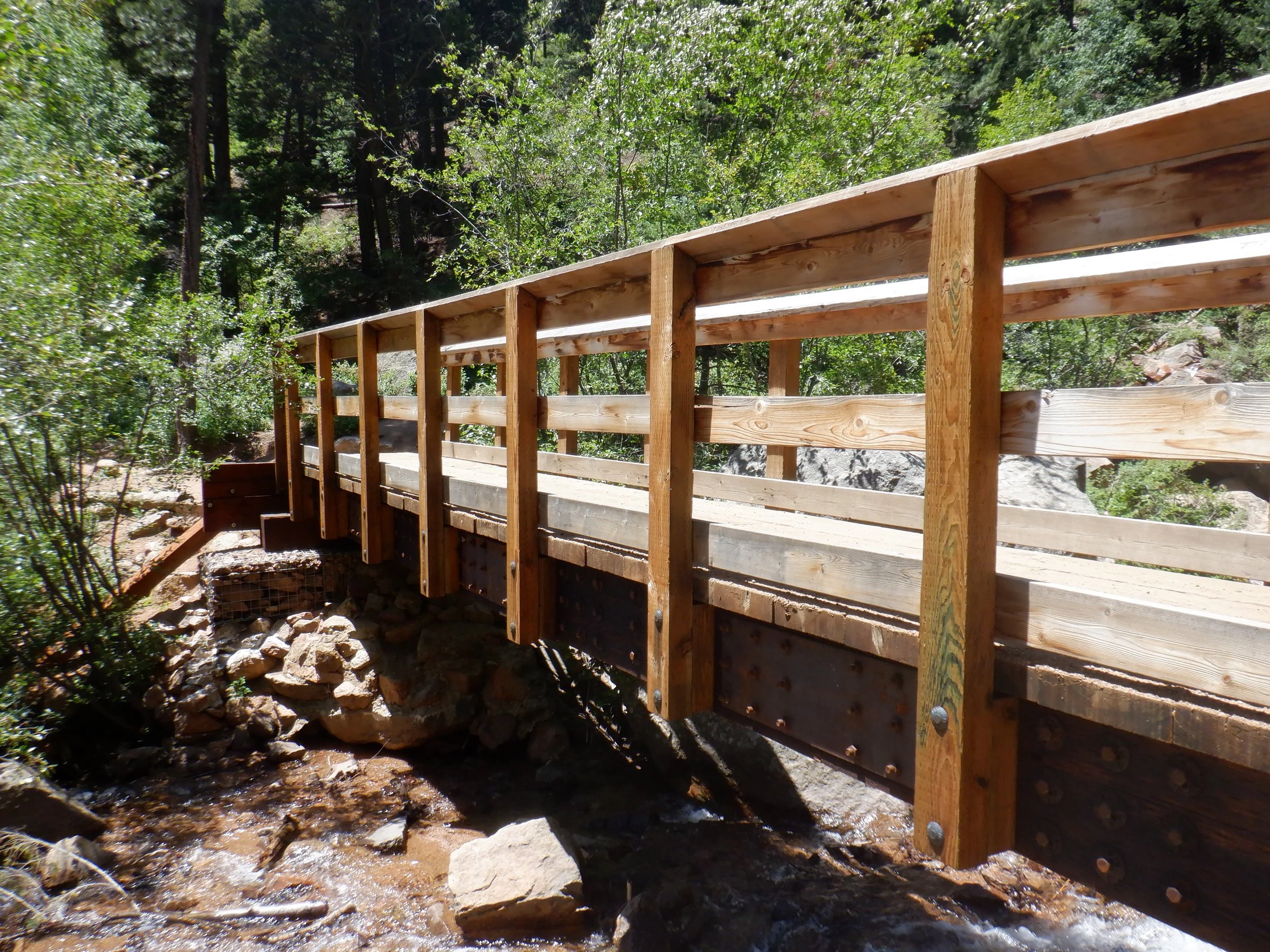 Wooden bridge over a creek in a forested area with trees and rocks.