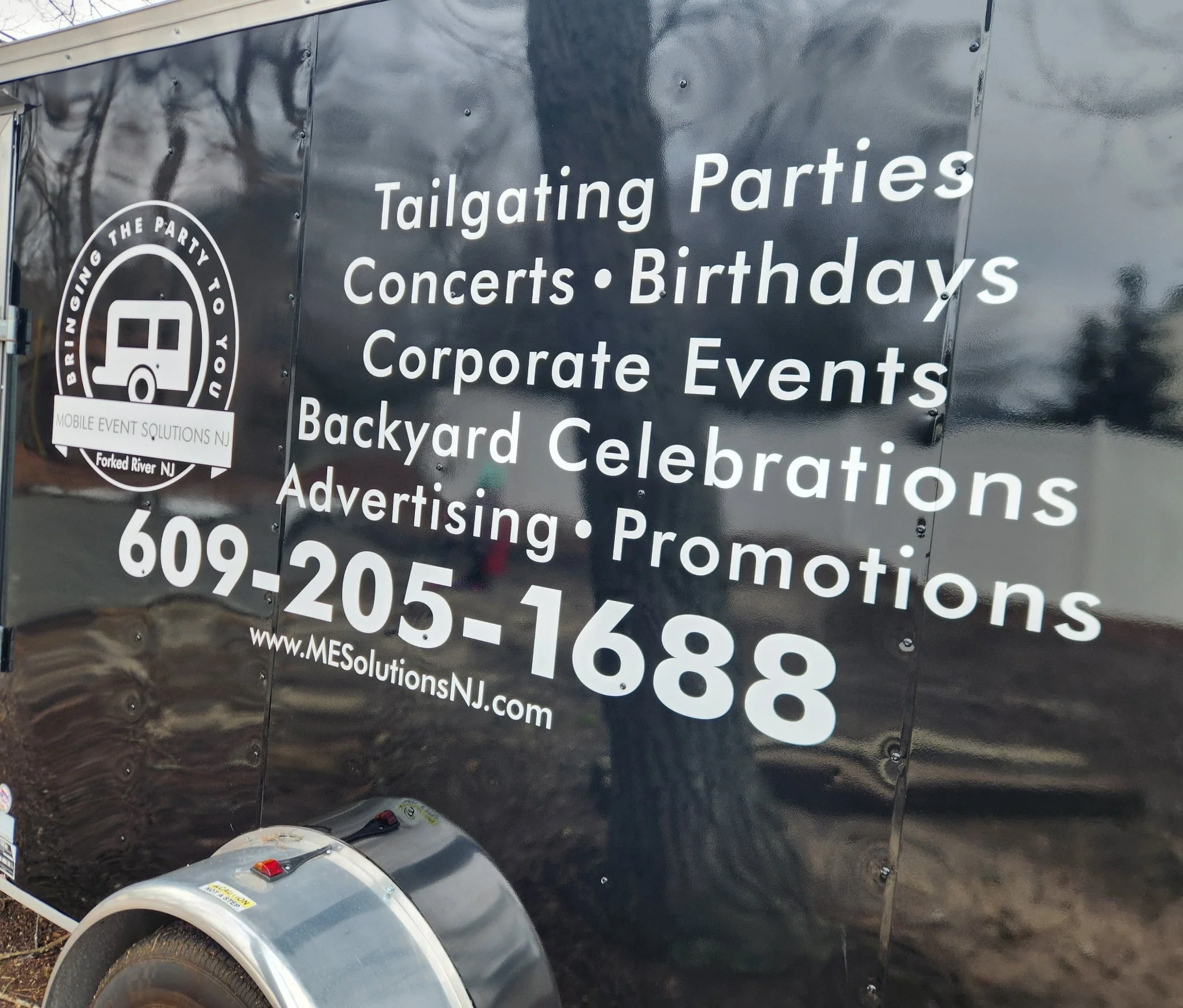 Black trailer advertising event services including tailgating parties, concerts, birthdays, corporate events, backyard celebrations, and promotions, with contact number 609-205-1688 and website www.MESolutionsNJ.com.