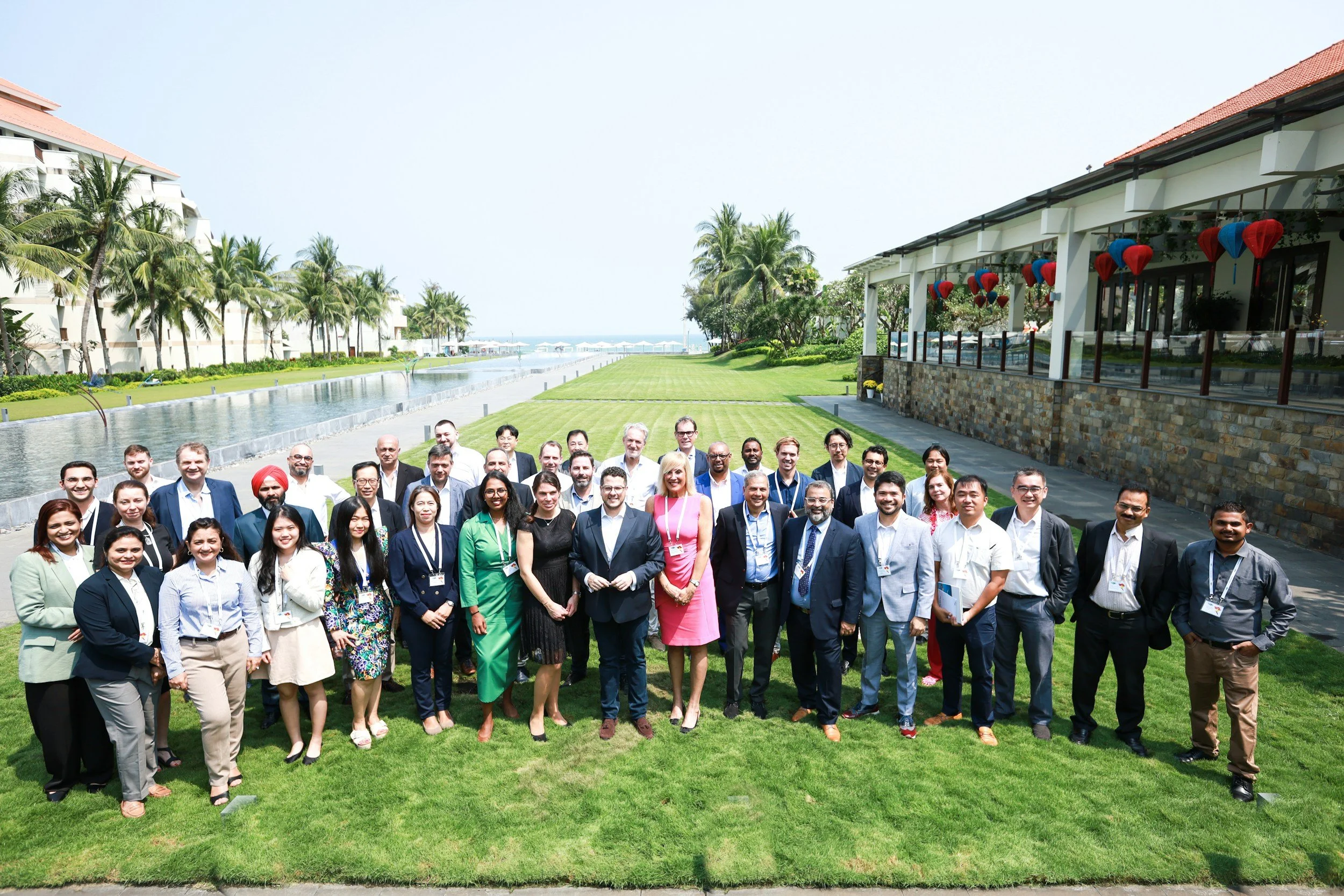 Group of diverse people in business attire standing on a lawn near a water feature at a tropical resort, with palm trees and a building decorated with red and blue lanterns in the background.