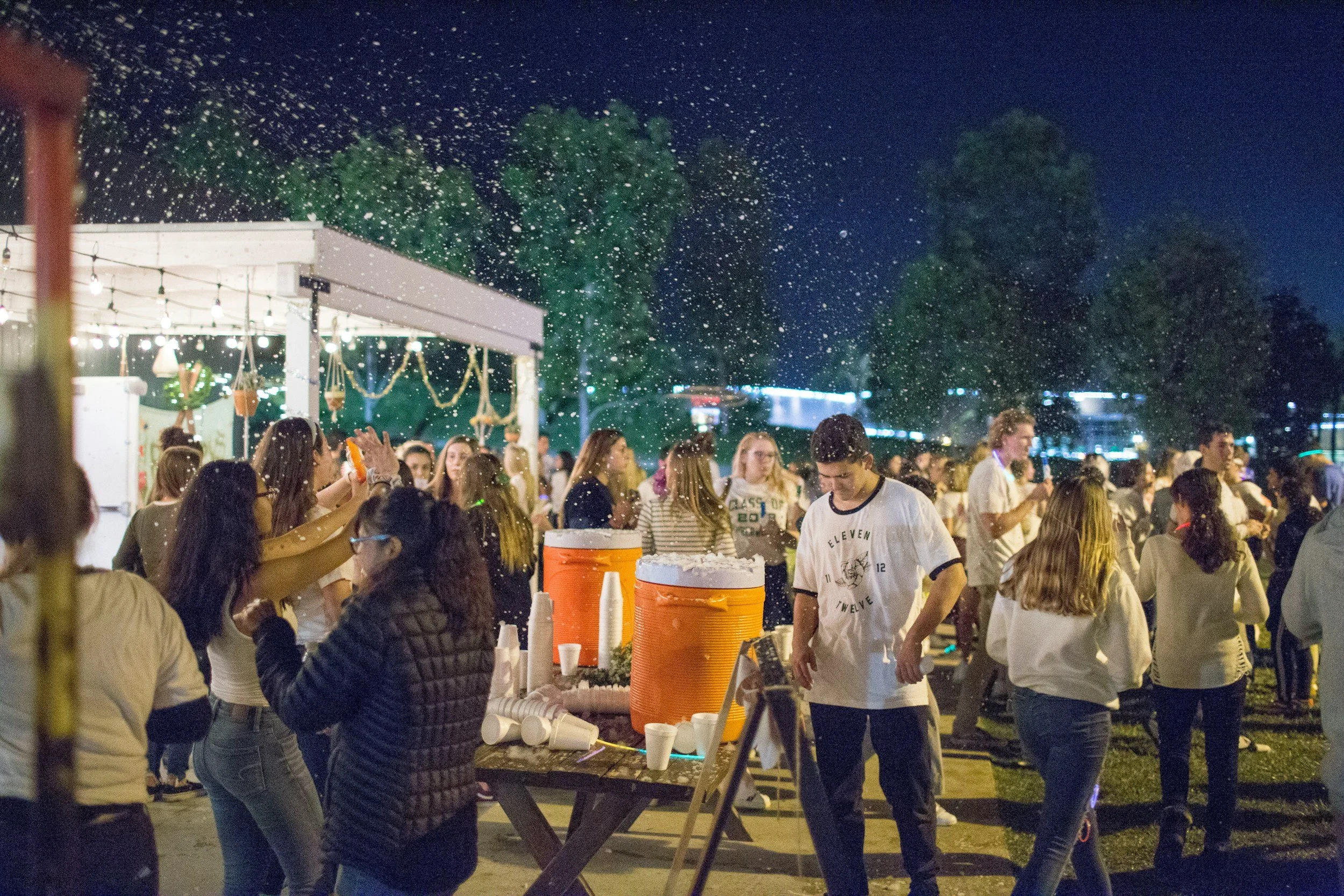 People socializing at an outdoor evening party, with snow falling and string lights hanging overhead. There are large orange coolers on a table, and some guests are holding cups.