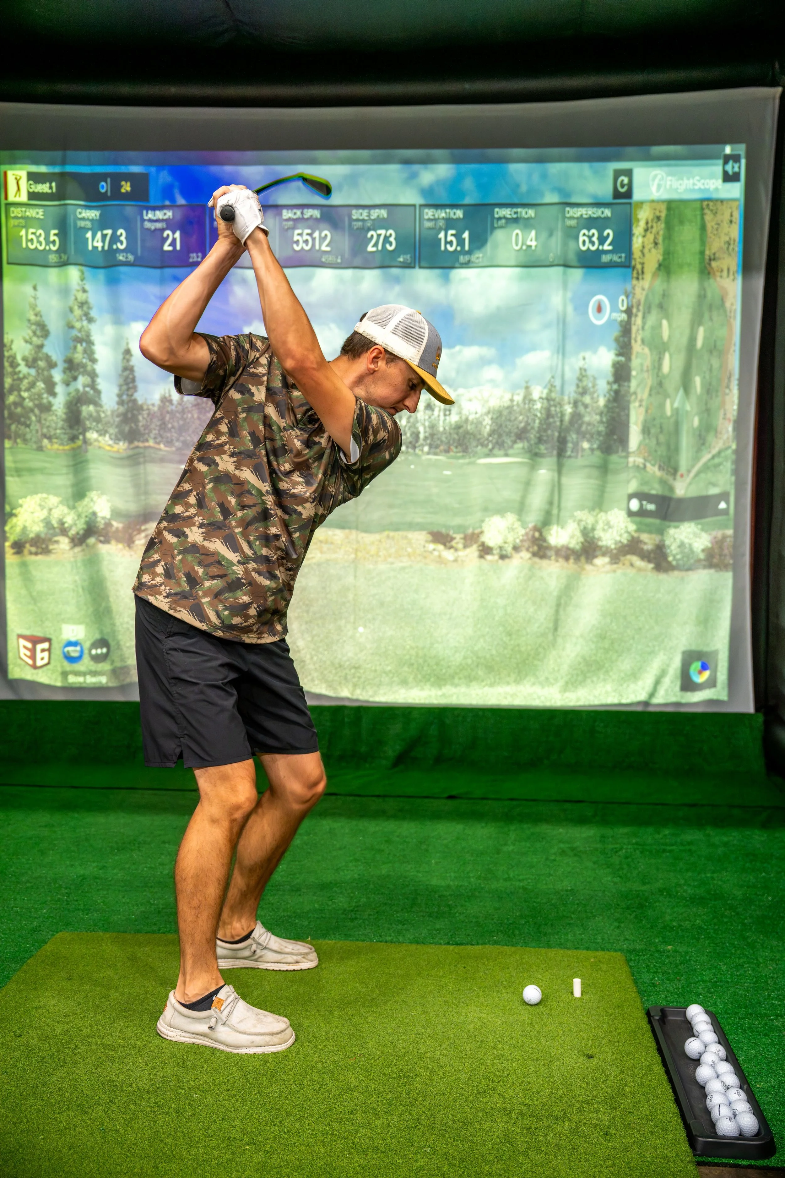 A man in casual clothes and a cap is practicing golf swings on an indoor golf simulator with a large digital screen showing golf course data and a map in the background.