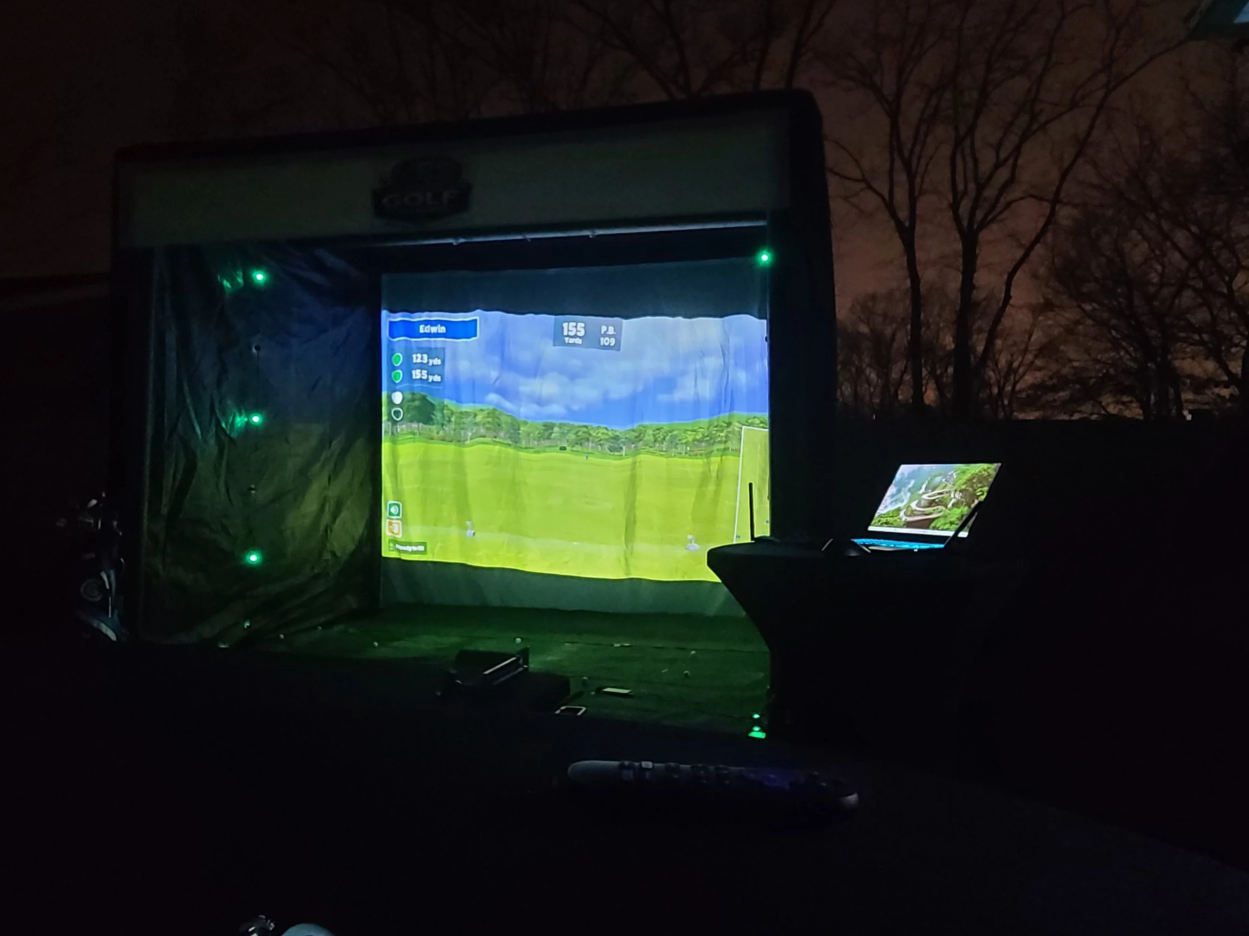Outdoor golf simulator setup at night with a large screen displaying a golf course and a laptop on the right side showing the same golf course.