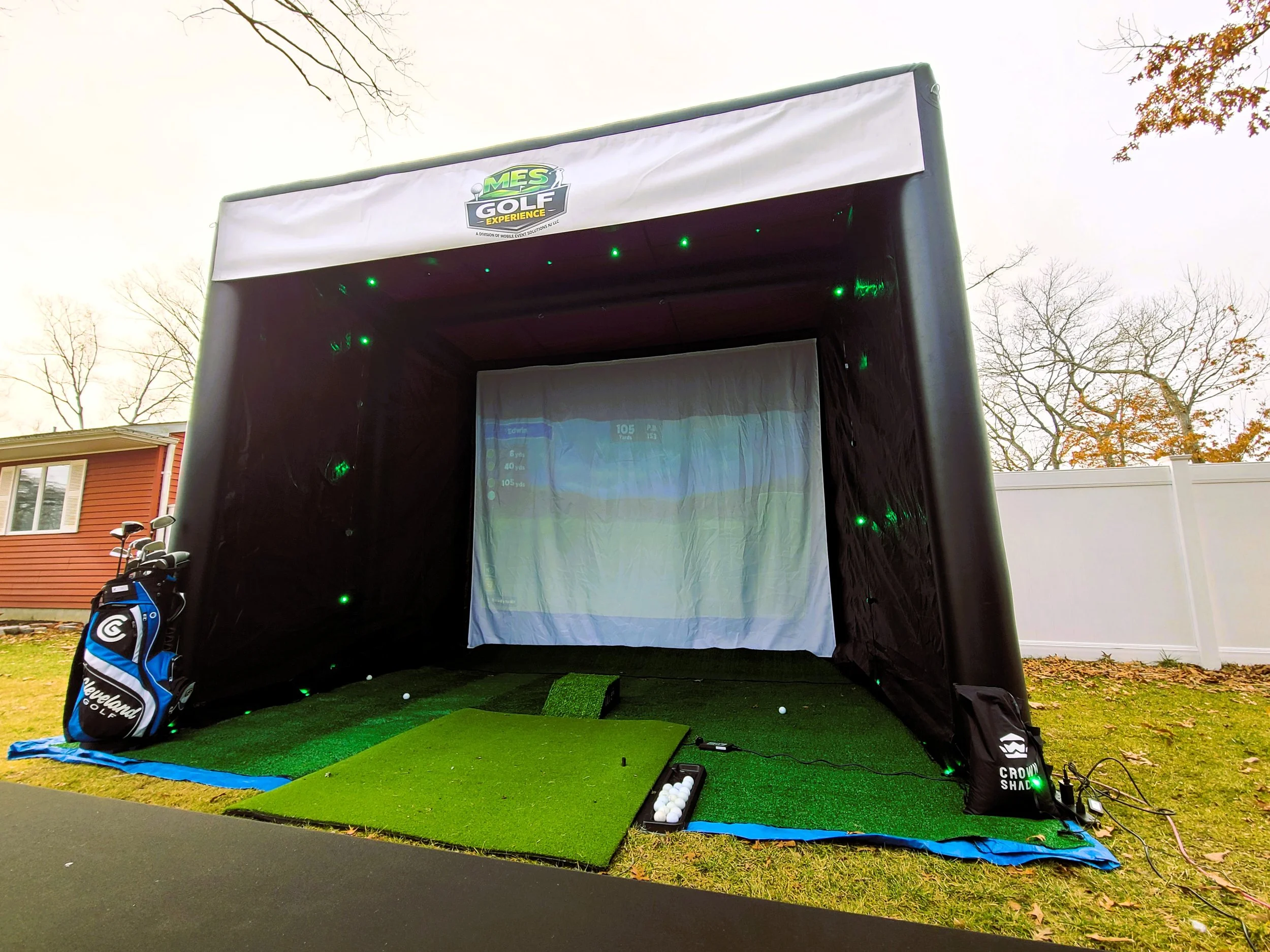 A golf simulator setup outdoors with a screen, artificial turf, golf clubs, and a golf bag, under a large black tent labeled 'MES Golf Experience'.