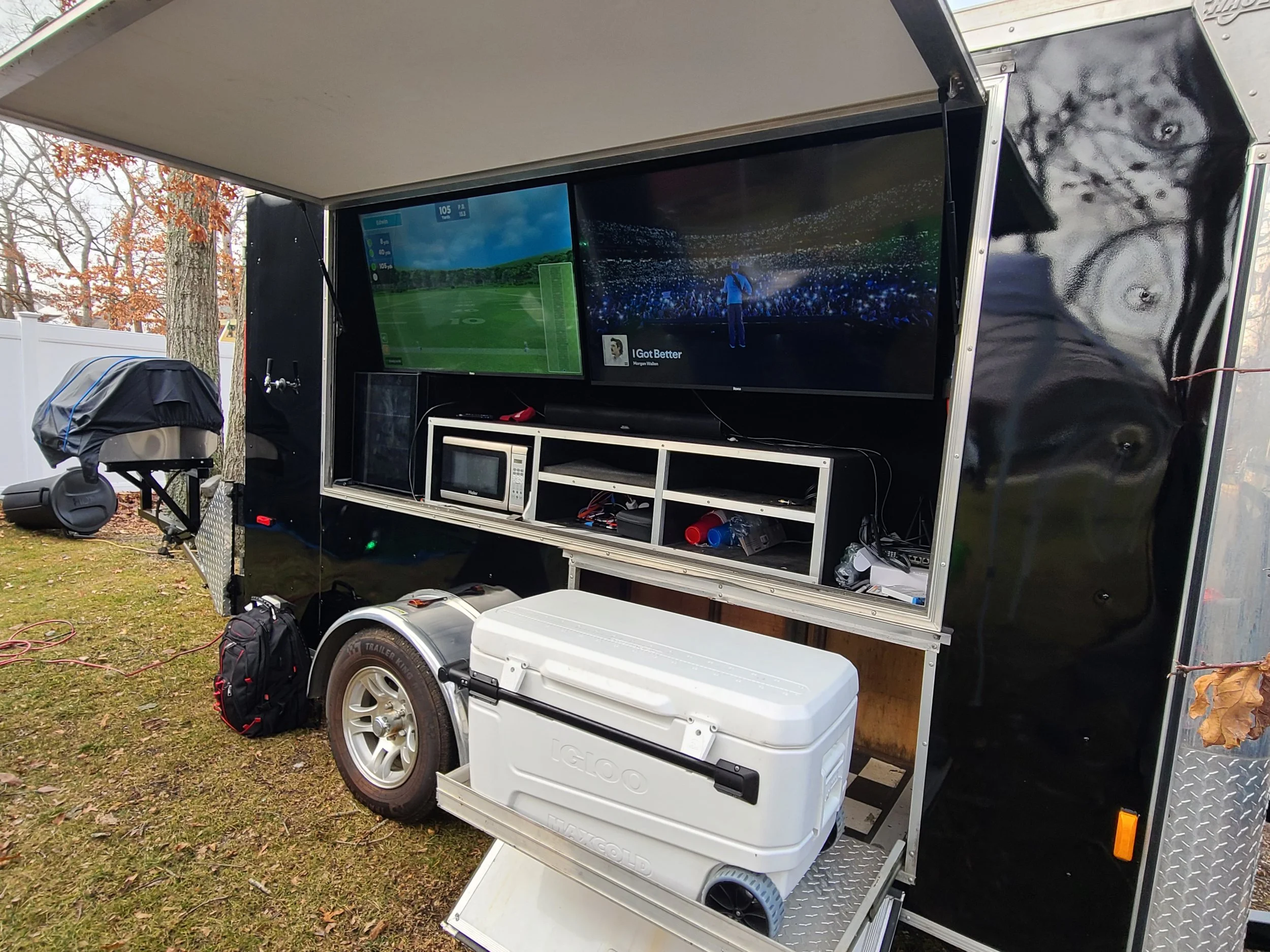 Mobile trailer setup with two large screens displaying a golf course and a concert stage, microwave, cooler, speaker, backpack, and other equipment outdoors on grassy ground.