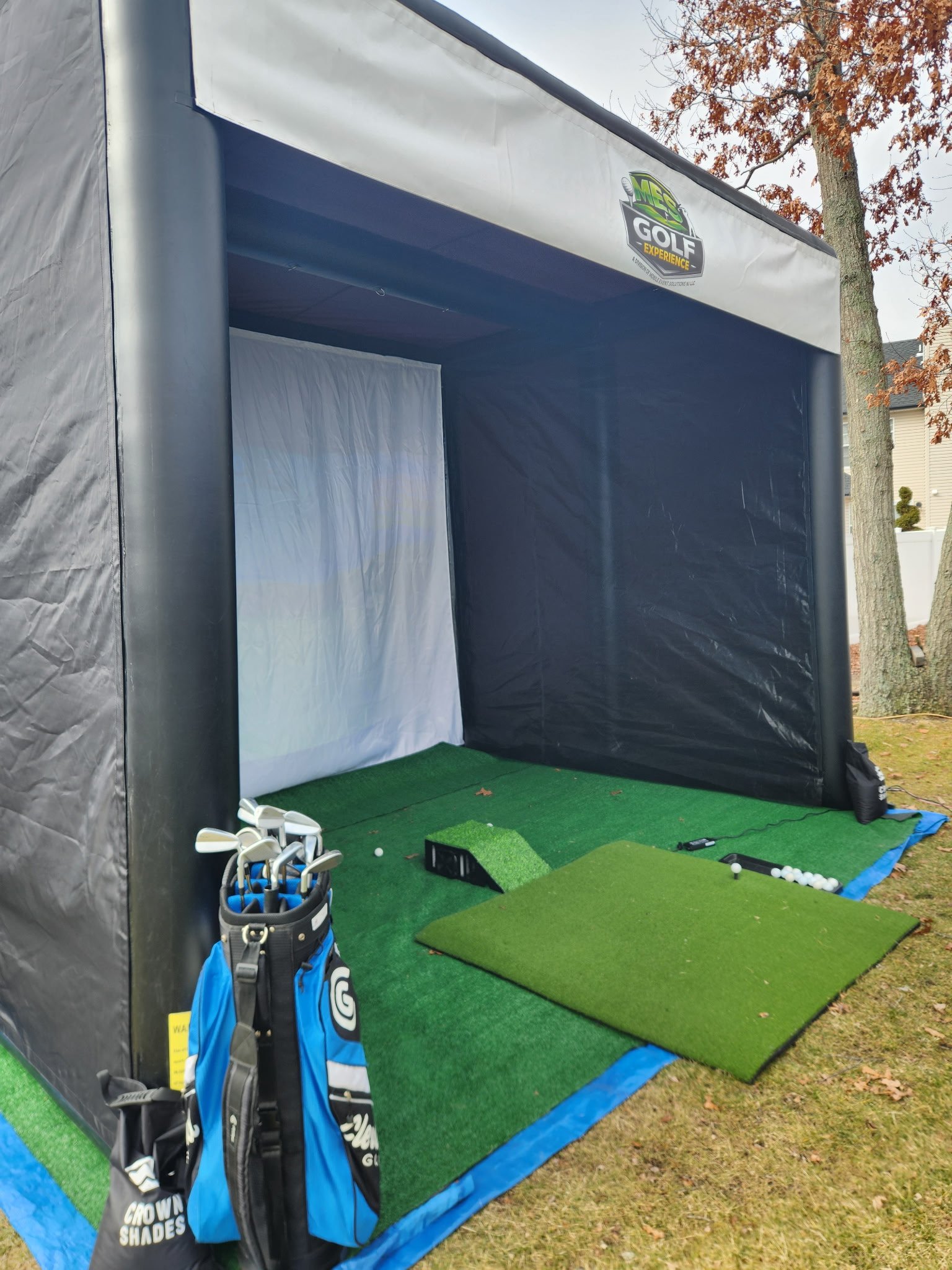 Mini indoor golf practice area with a green putting surface, golf clubs in a bag, and golf balls, set up outdoors with trees and a residential house in the background.