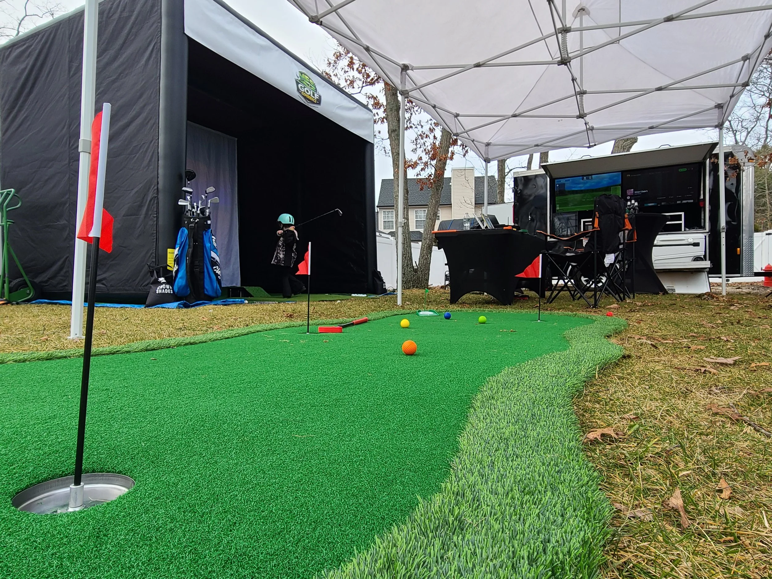 A portable golf simulator setup with a small putting green, flags, colorful golf balls, and a child wearing a helmet practicing putting. There are tables and screens inside a mobile trailer, shaded under a canopy, with golf equipment nearby and house