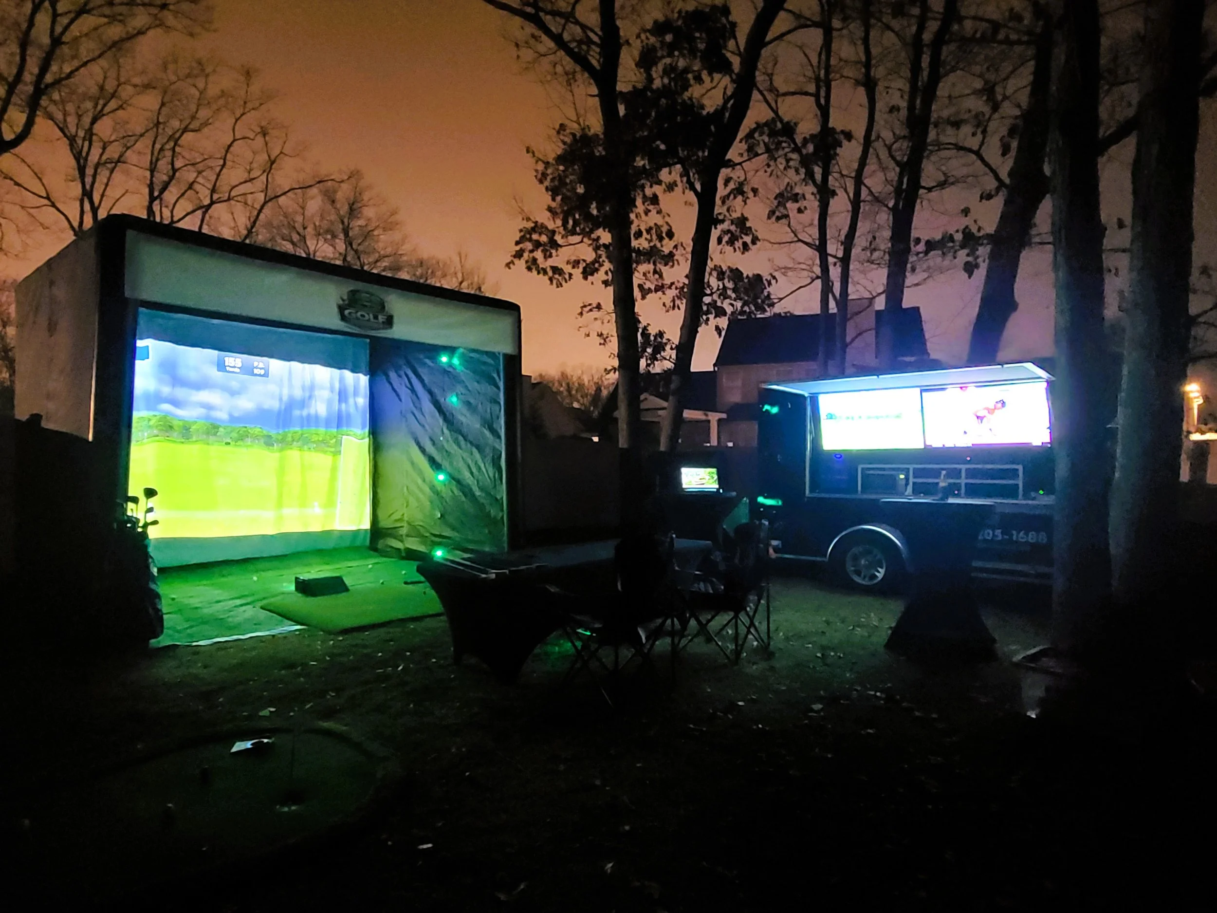 Outdoor miniature golf course illuminated at night with electronic screens showing animated graphics, surrounded by trees and chairs.
