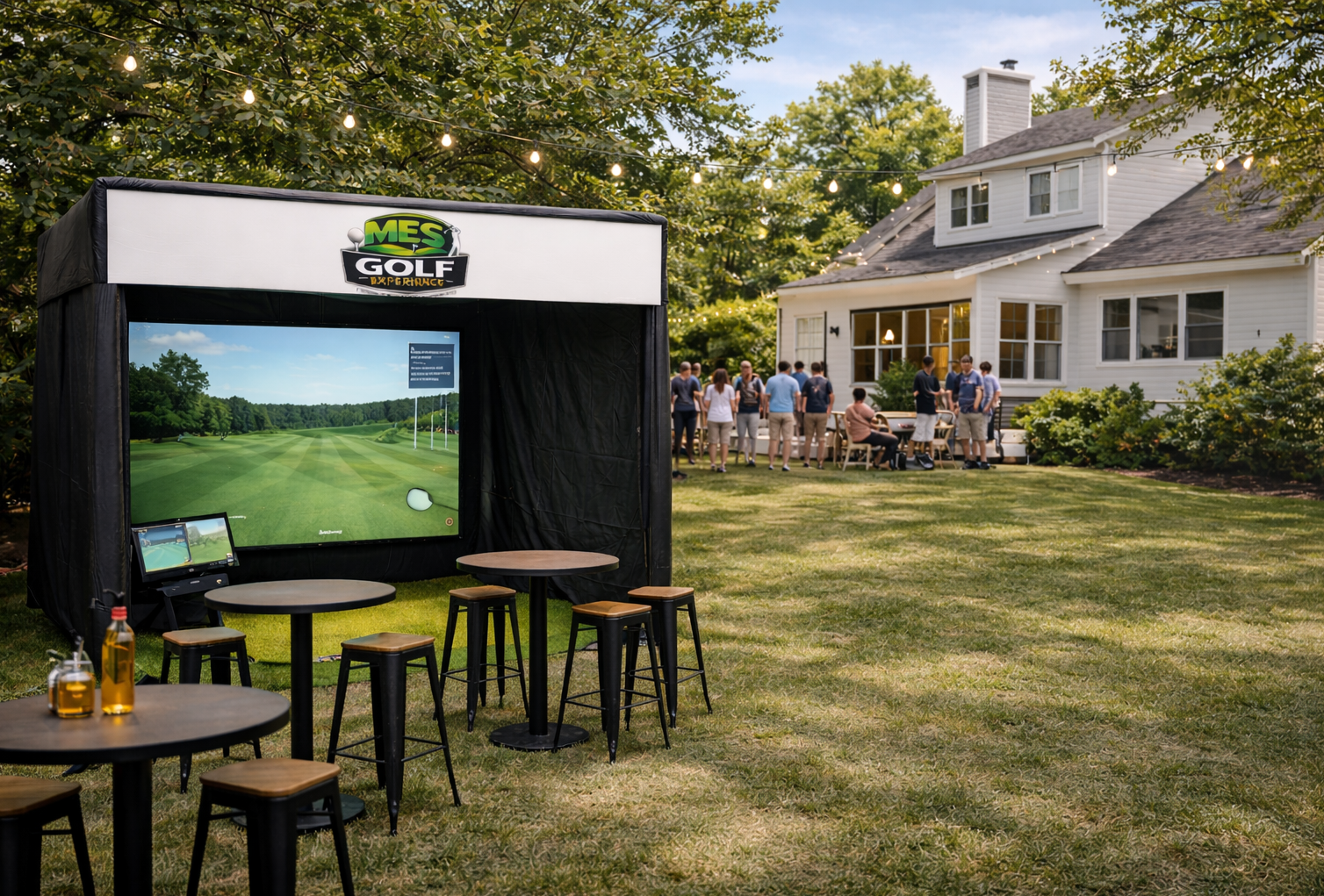 Outdoor setting with a golf simulation screen, tables, and chairs in front of a house with a group of people socializing on the lawn, surrounded by trees and string lights.